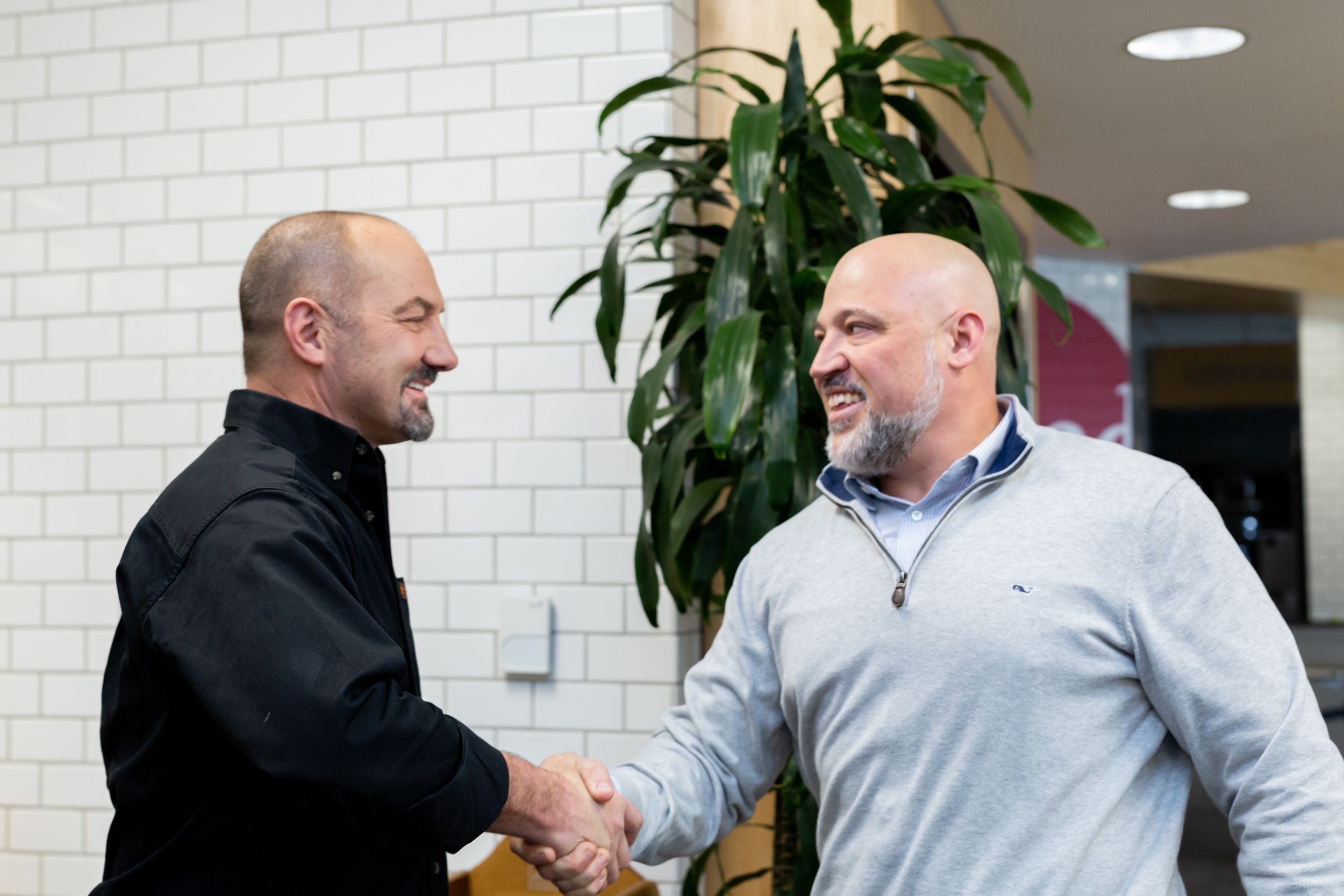Two men shake hands in a professional setting with white brick wall and potted plant in background.