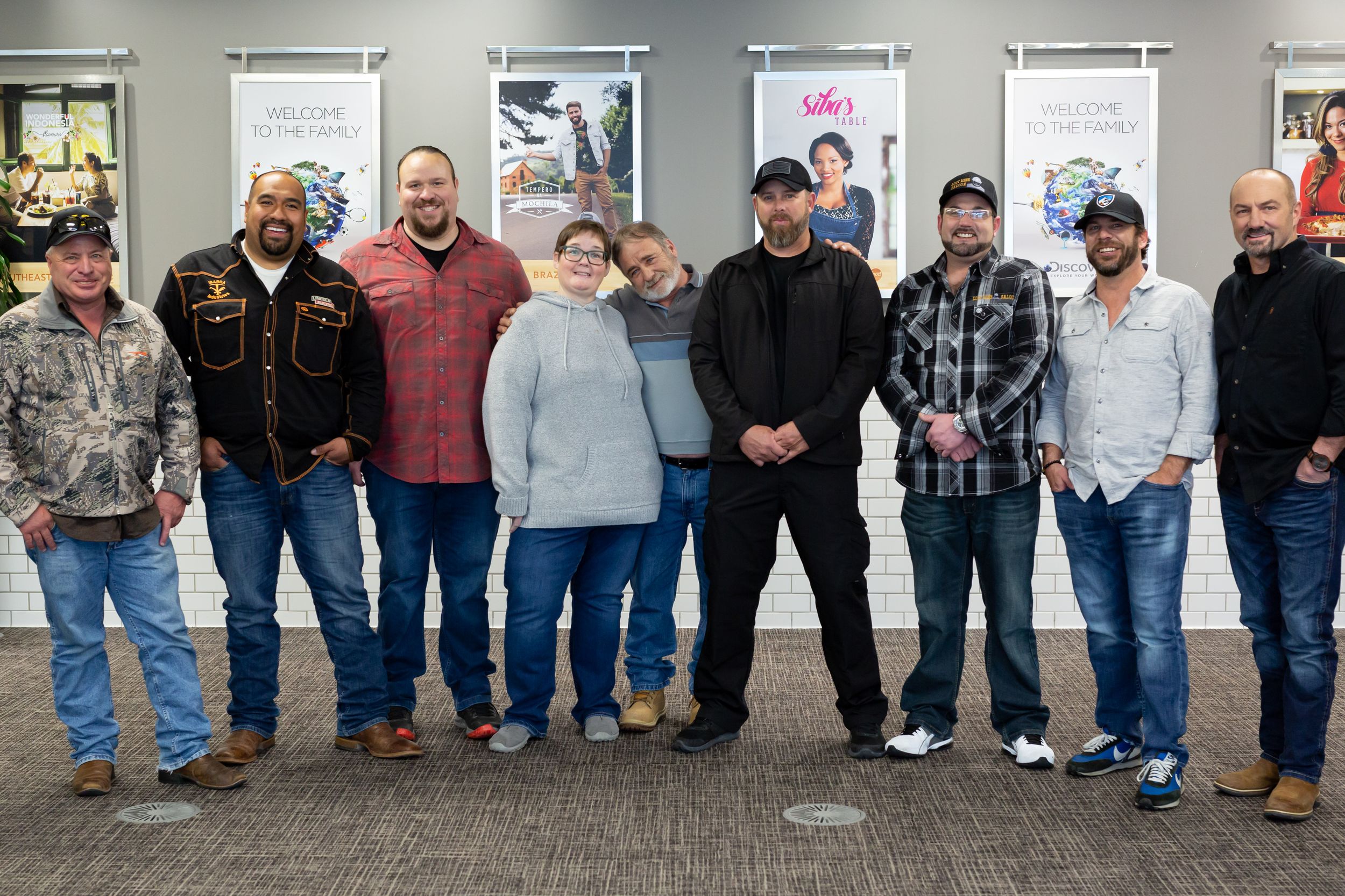 A group of people stand together in a line against a wall with framed pictures, dressed in casual work attire.