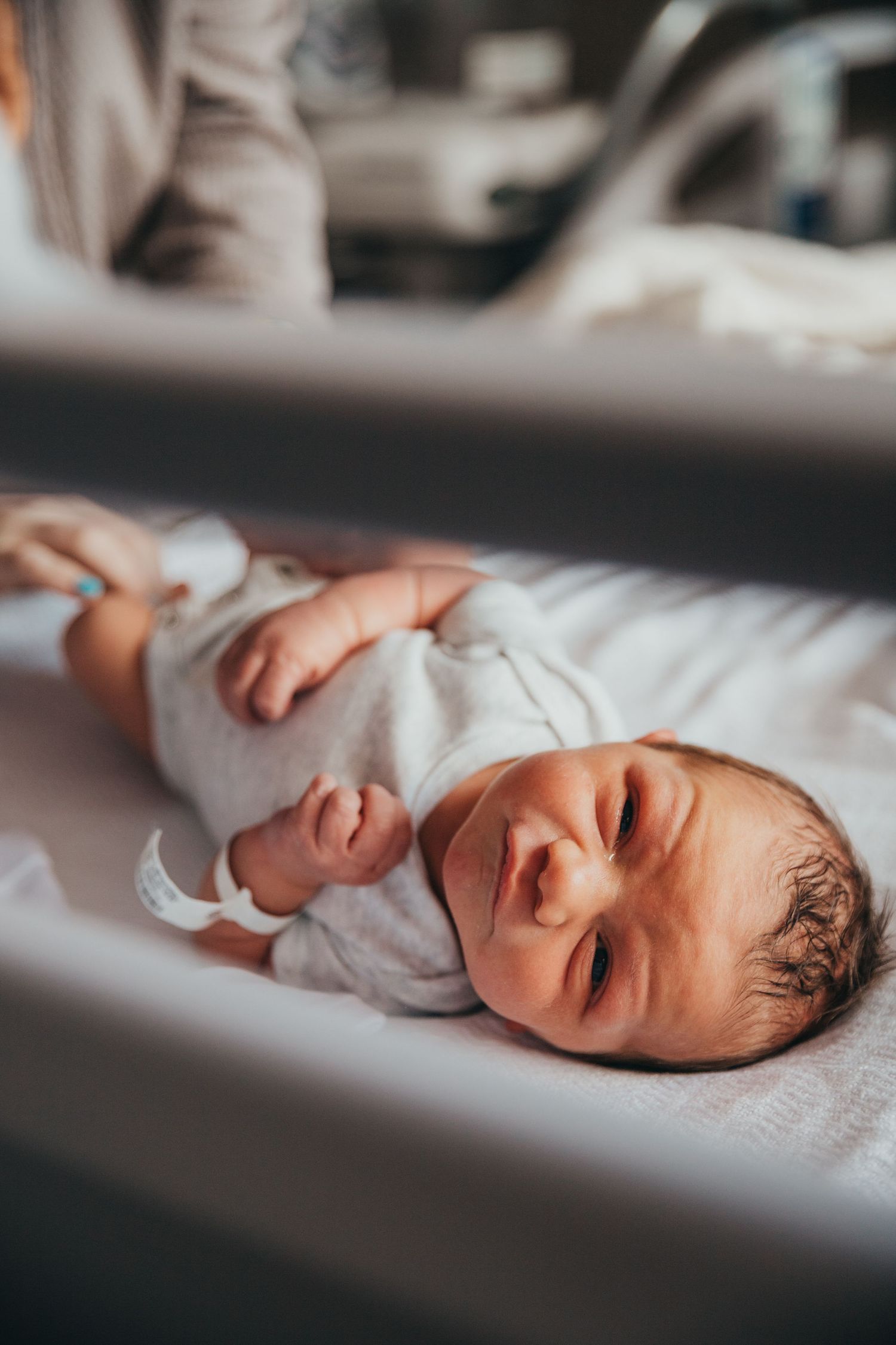 Newborn baby lying in a hospital bassinet wearing white clothing, photographed from above in soft lighting.