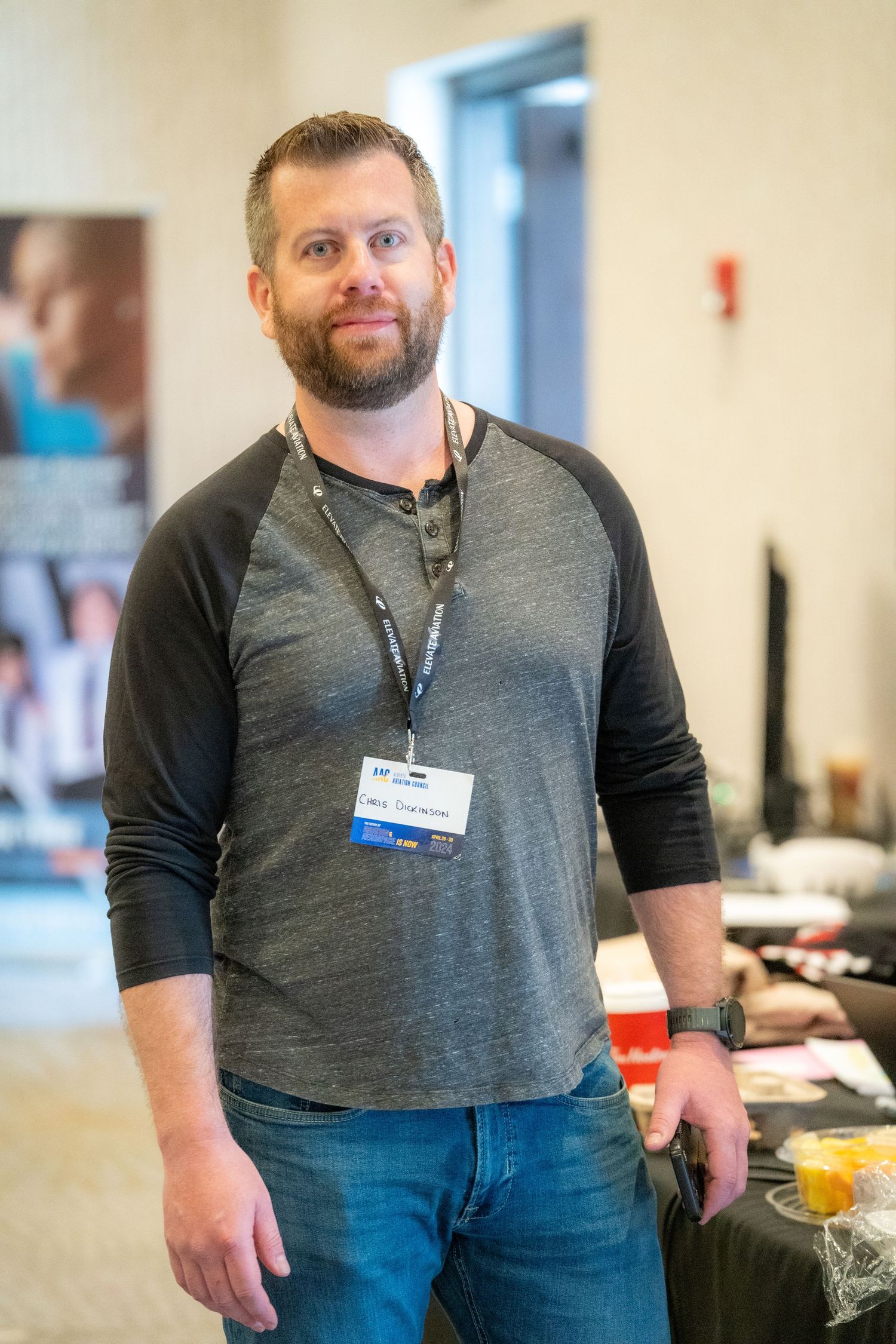 A person wearing a gray henley shirt and jeans stands in an office hallway with a name badge lanyard.