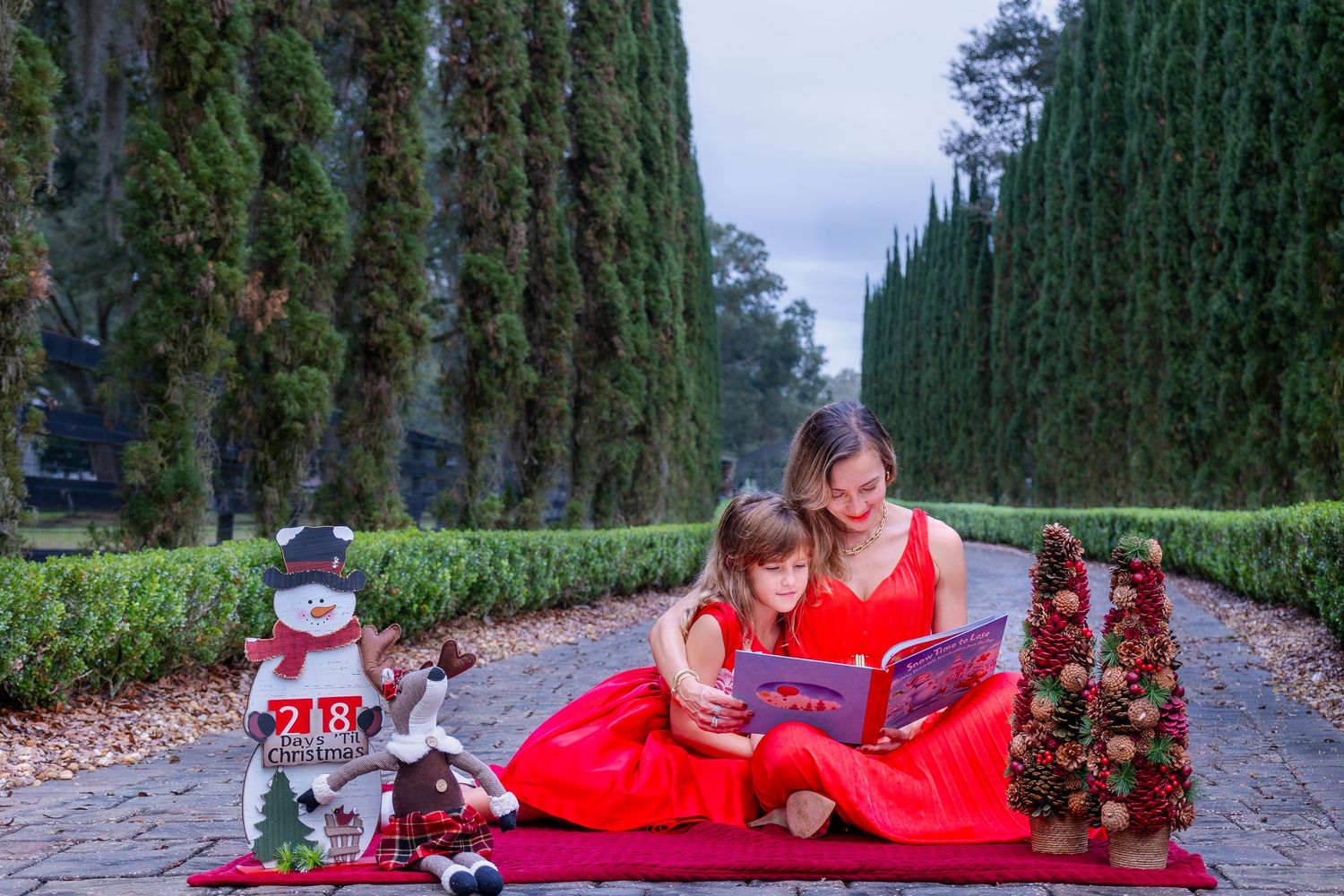 Reading together on a red blanket surrounded by cypress trees and holiday decorations on a winter path.