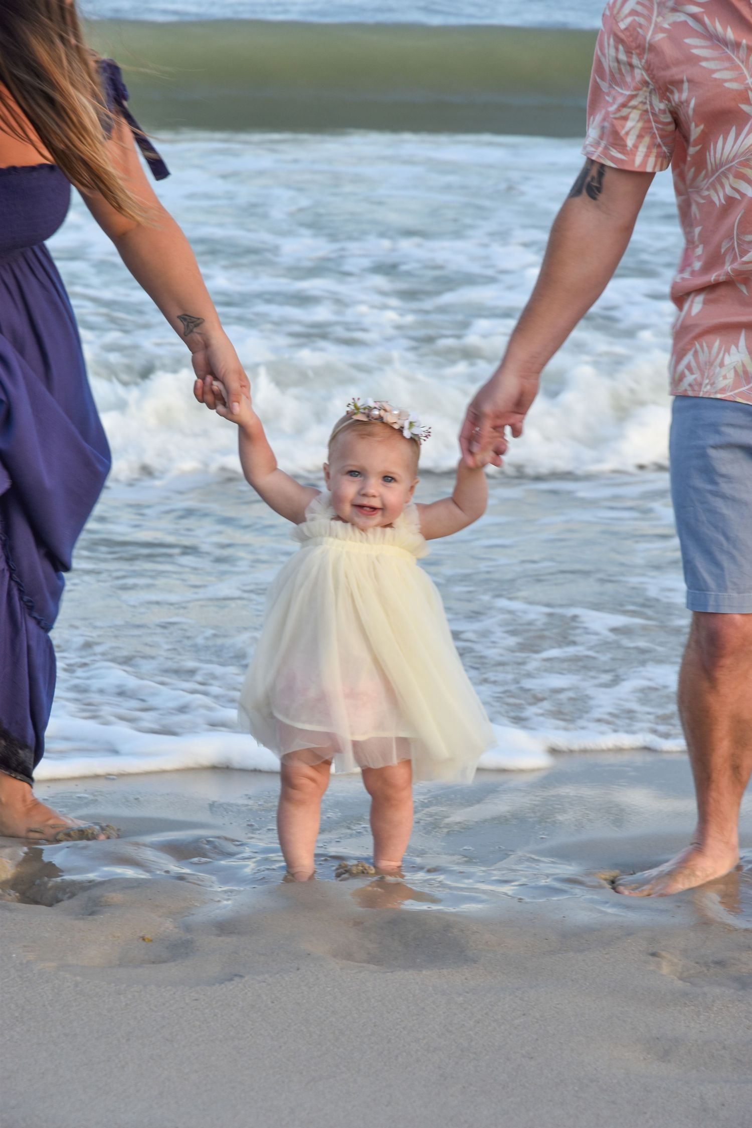 A toddler in a tulle dress plays in the ocean waves while holding hands with parents.