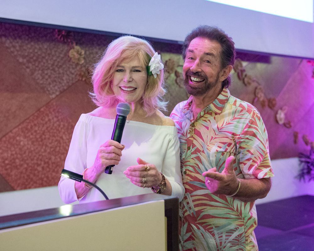 Two presenters on stage sharing a microphone during an event with colorful lighting.