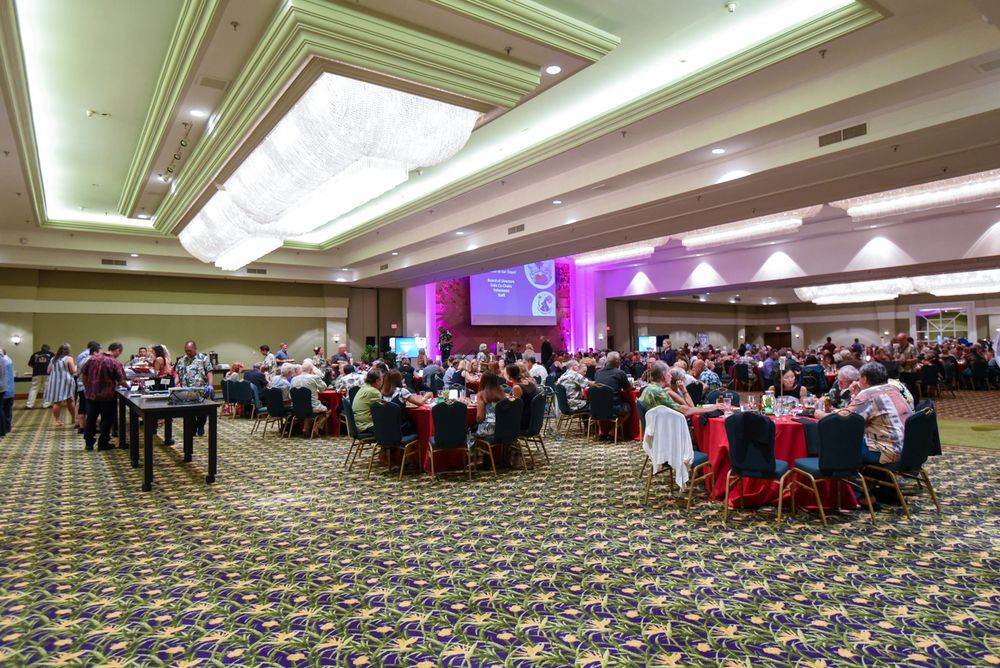 Large banquet hall with round tables and attendees at an event with purple lighting and patterned carpet.