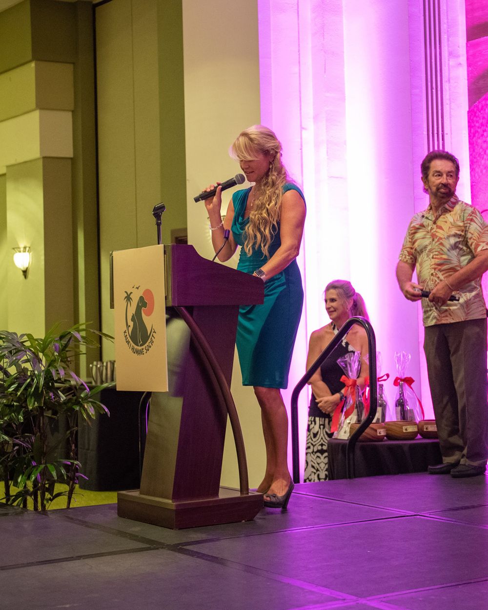 Woman in blue dress speaking at podium during formal event with pink-lit backdrop.