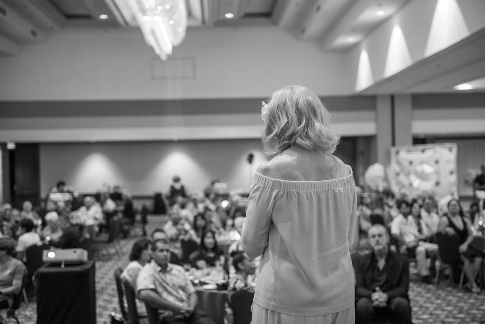 A speaker addresses a large audience in a hotel conference room from stage, shown from behind.