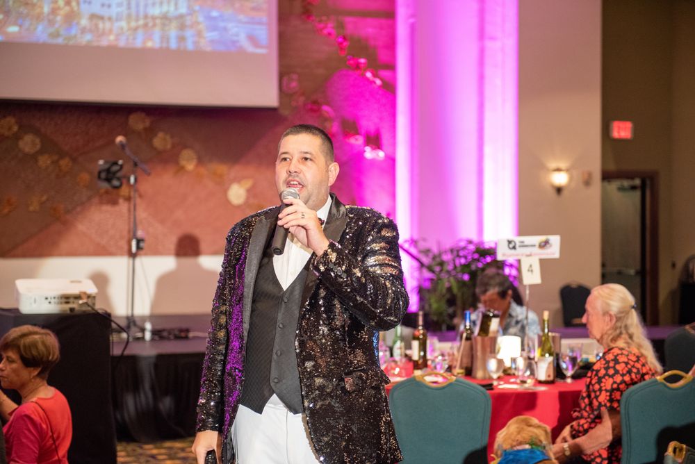 Person in sparkly jacket speaking into microphone during formal event with pink lighting.