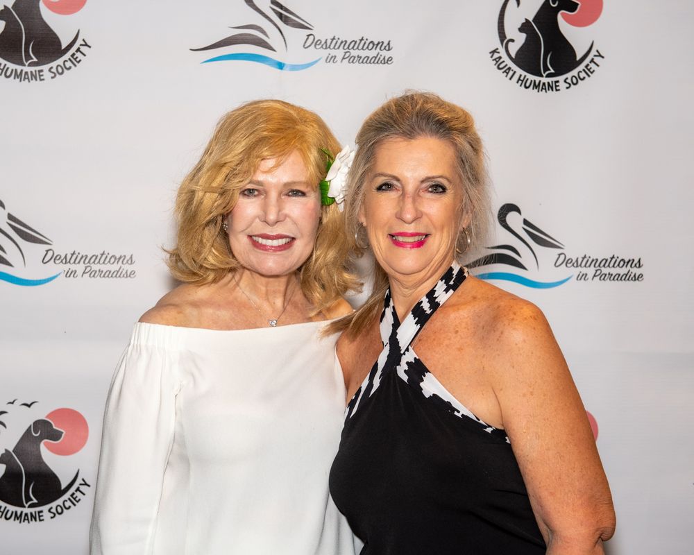 Two women posing at a charity event backdrop with palm tree and swimmer logos.