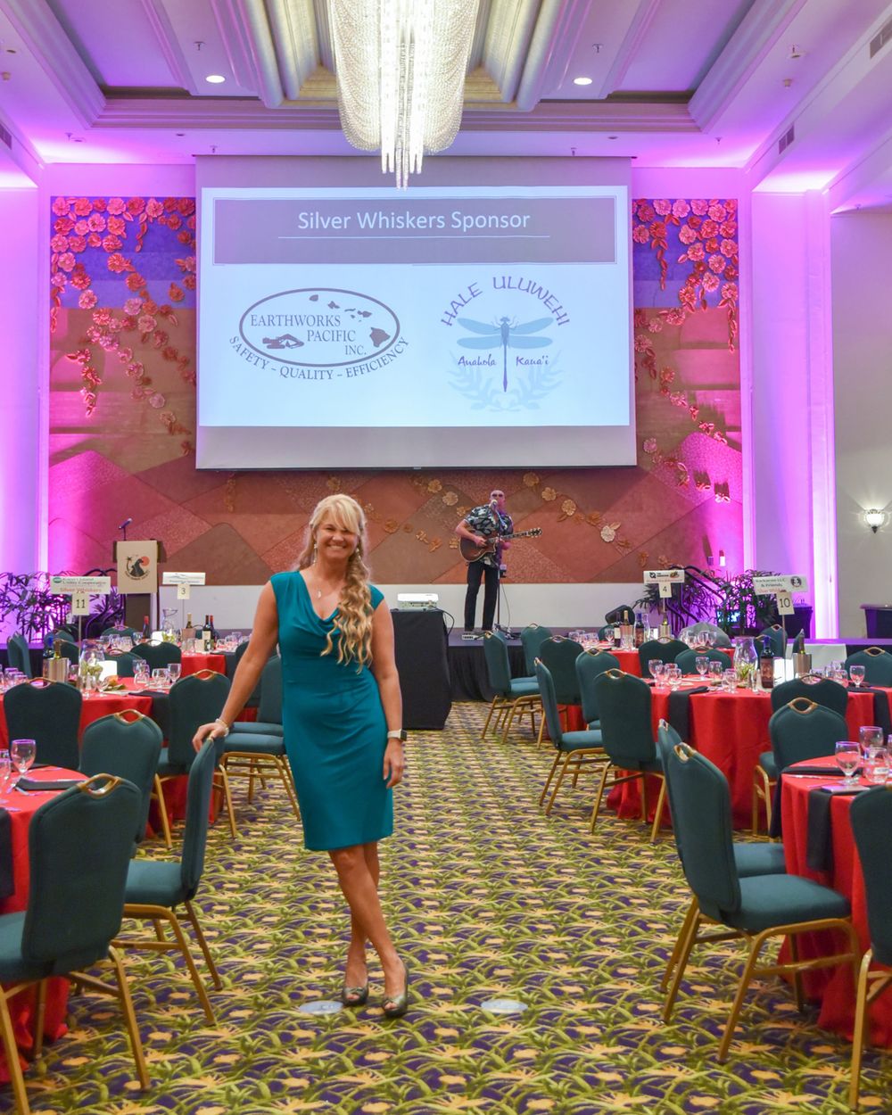 Woman in teal dress standing in decorated ballroom with presentation screen and red table settings.