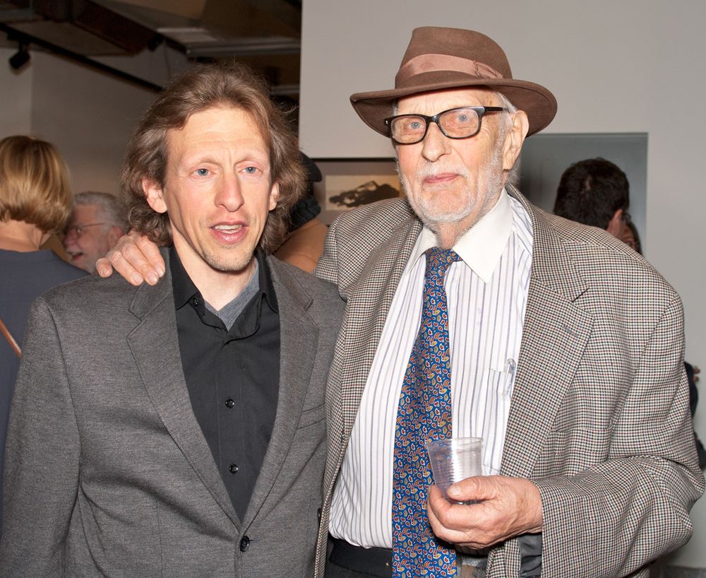 Two men at formal event pose together, one wearing hat and glasses.