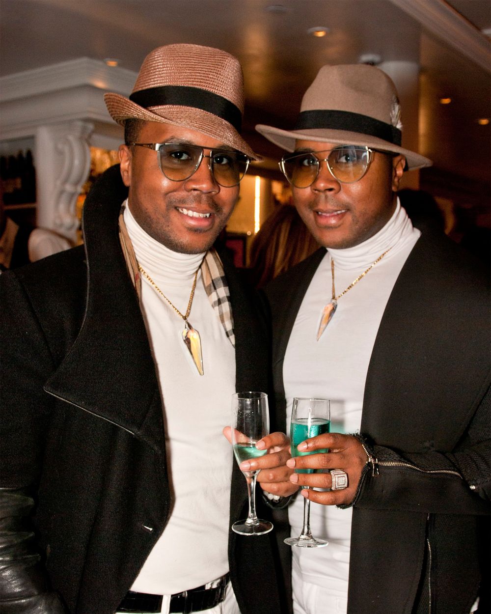 Two stylish men in matching fedoras and formal wear holding champagne glasses.