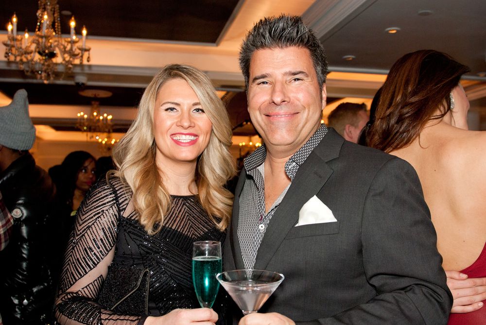 Couple in formal wear holding drinks at evening event with chandelier in background.