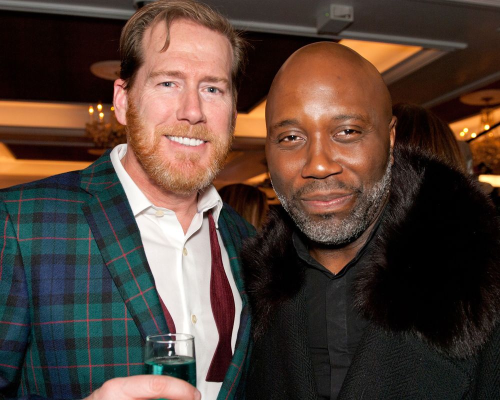 Man in plaid jacket and man in black outfit pose together at indoor event.