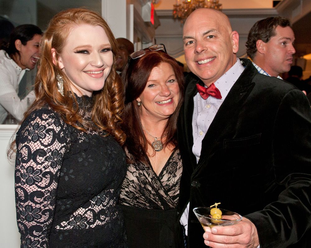 Three people in formal attire pose together at an evening event, with one wearing a distinctive red bowtie.