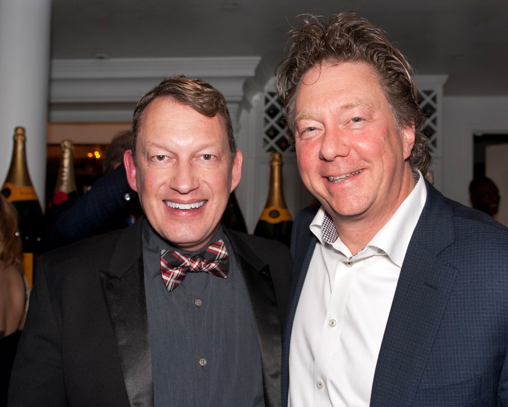Two men in formal attire smile warmly at a social gathering, one wearing a bowtie and the other in a blazer.