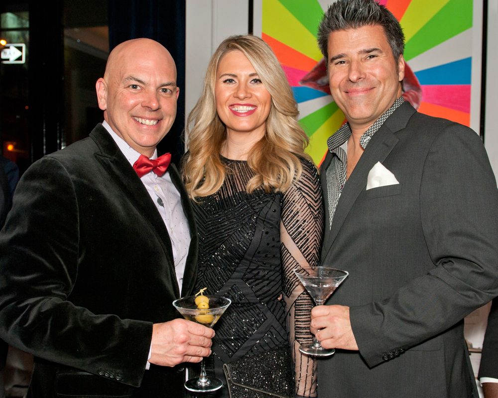 Three people smile at a party in front of a colorful geometric wall display while holding cocktails.