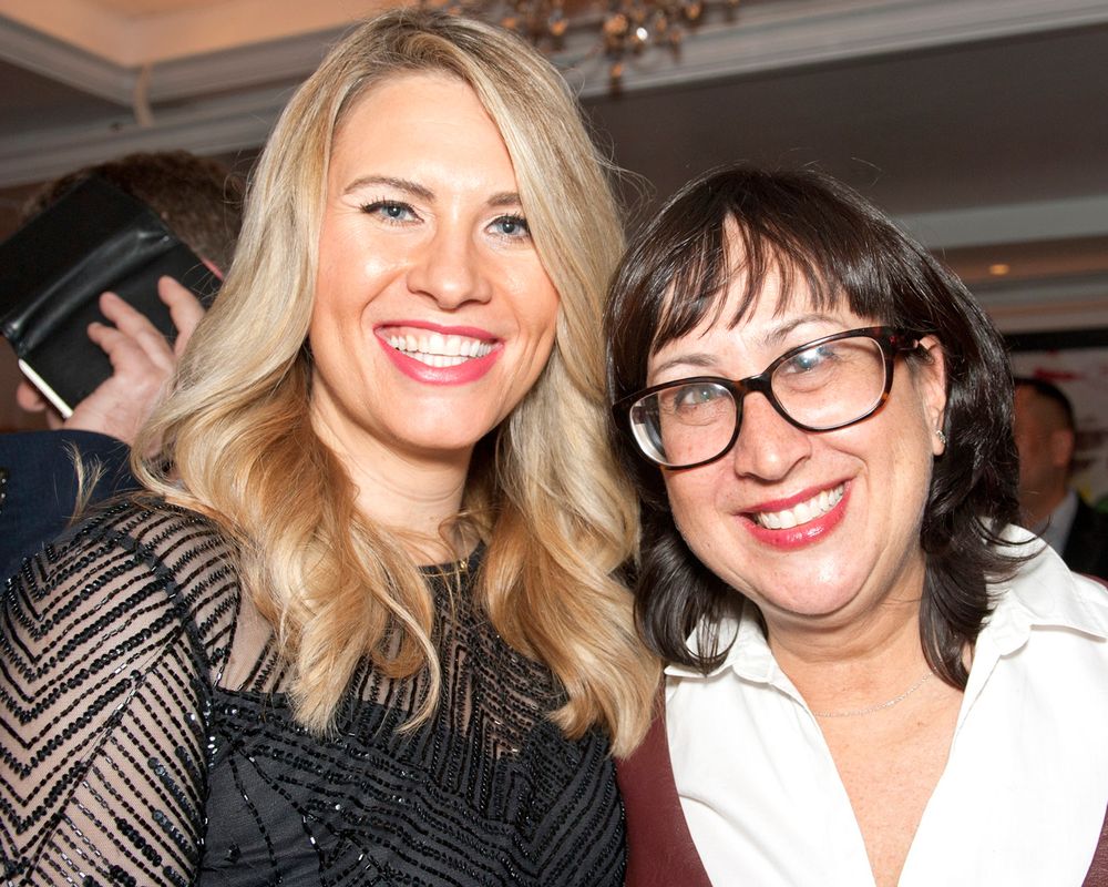 Two people pose together at an event, one wearing glasses and both smiling brightly at the camera.