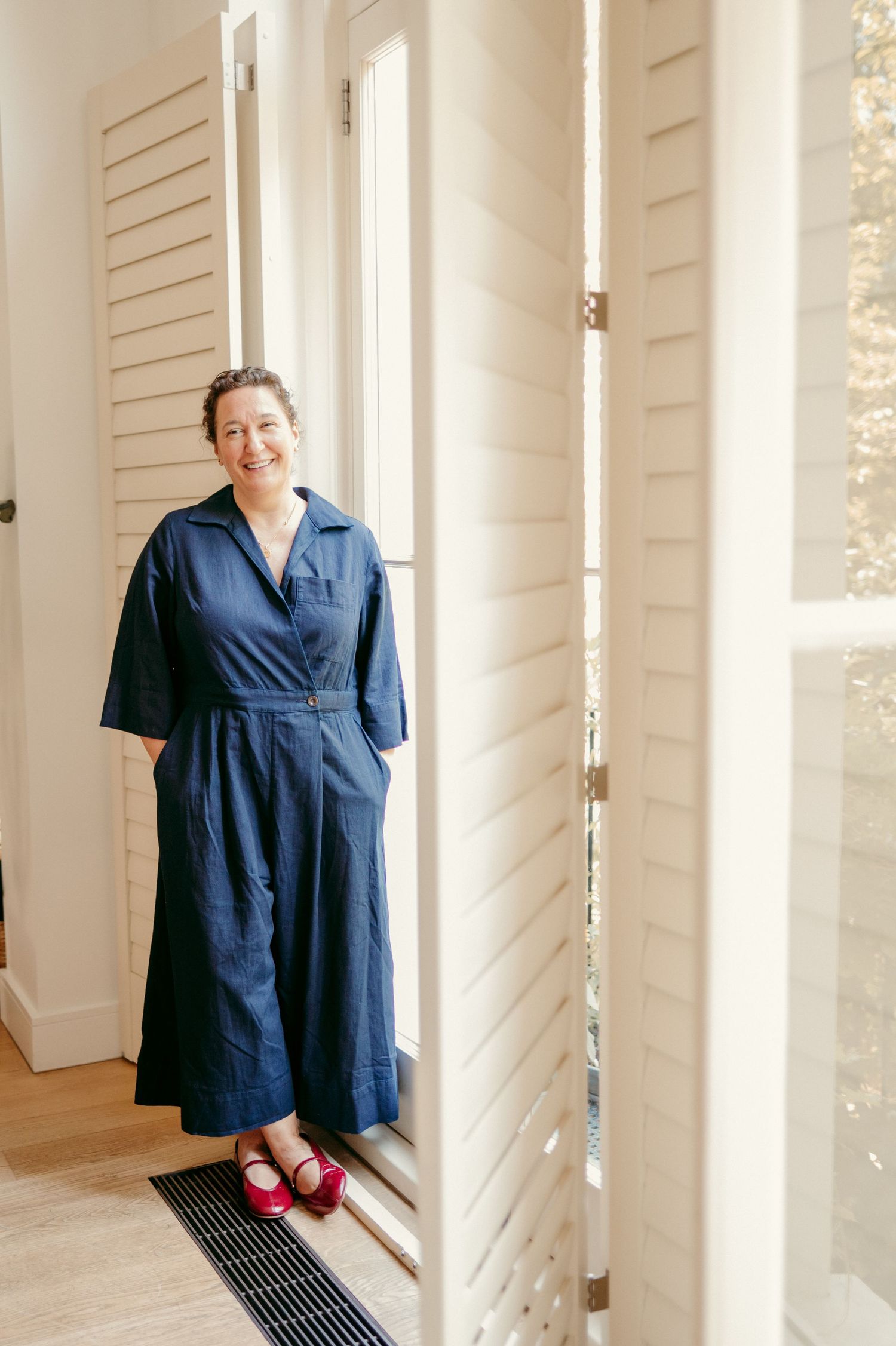 Woman wearing a navy blue wrap dress and red shoes stands in a bright white room.