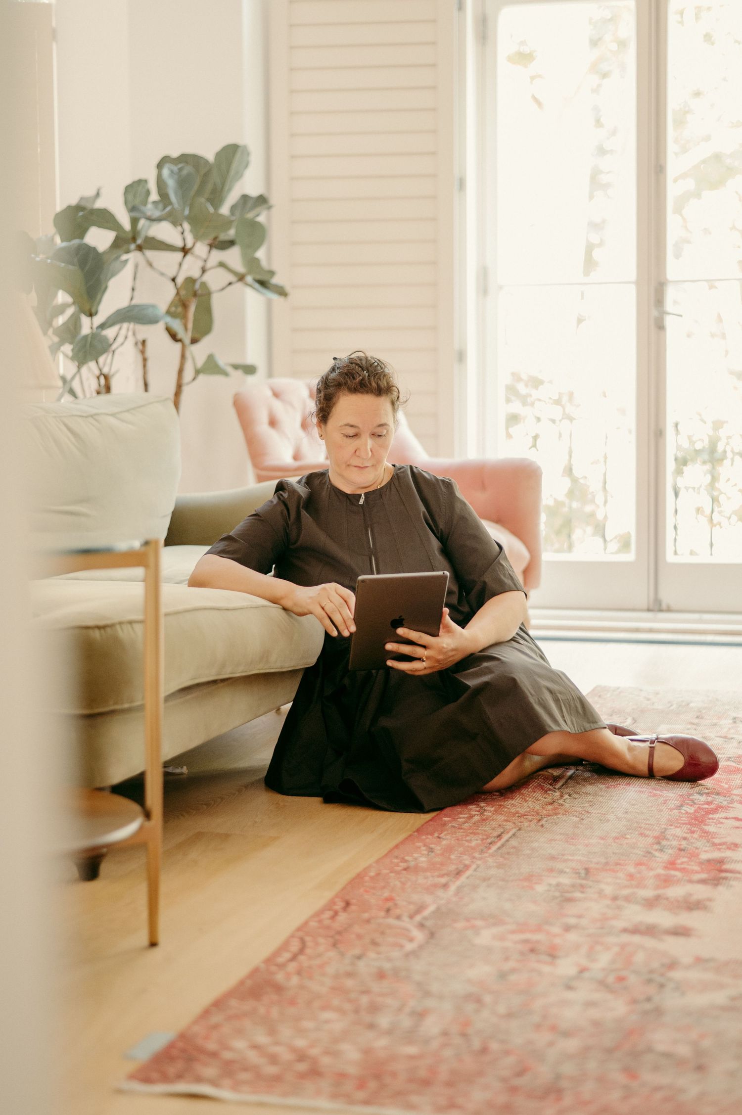 Someone relaxes on the floor against a couch while reading a tablet in a naturally lit room with plants.