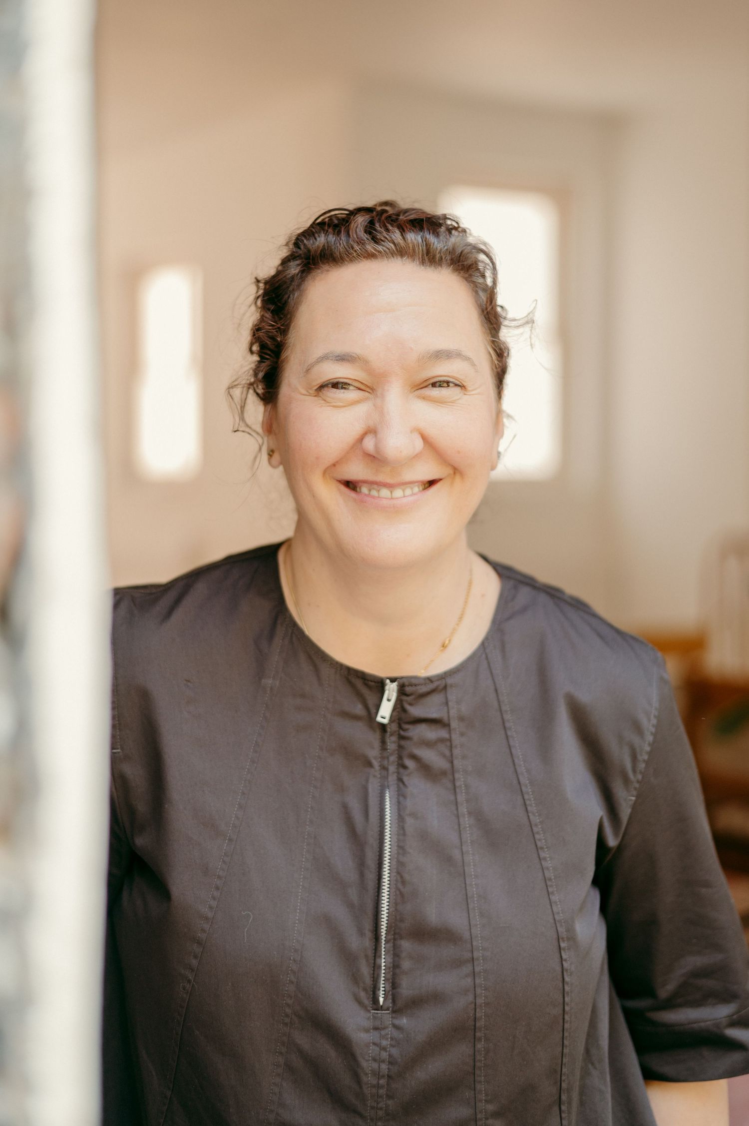 A person in a black zippered top smiles warmly at the camera in a sun-filled room.