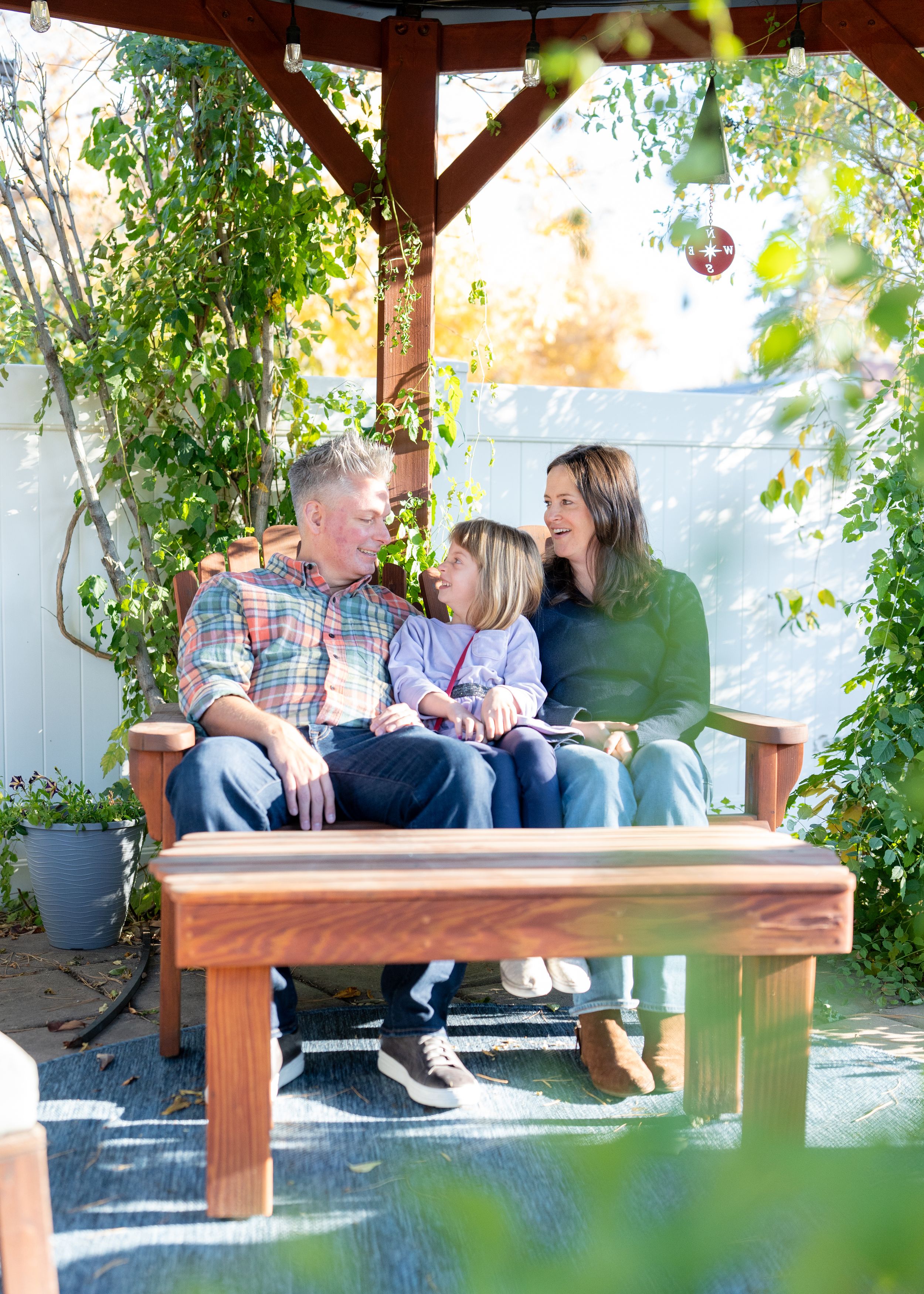 Family relaxes together on wooden bench in garden pergola.