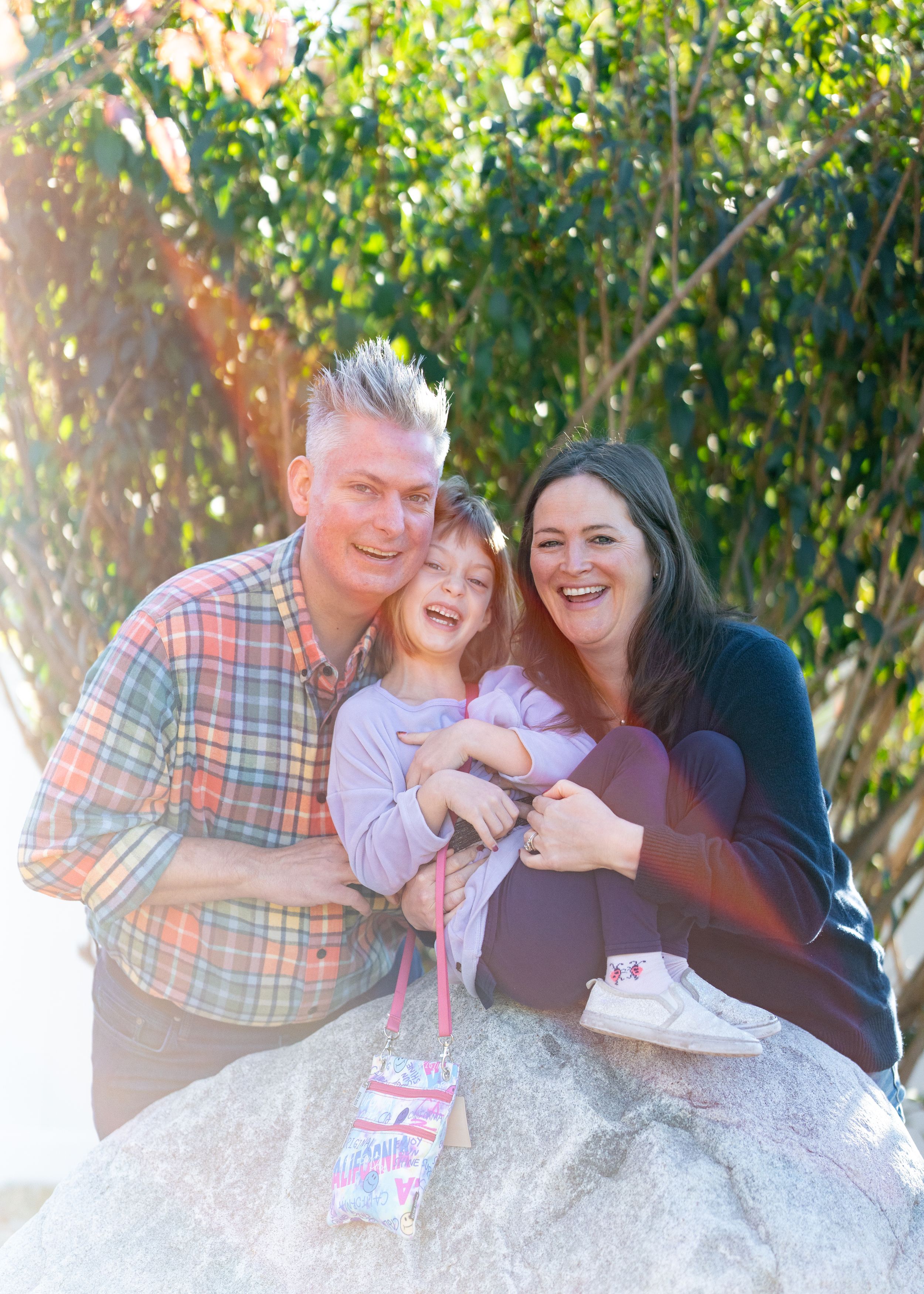 Family shares joyful moment together while sitting on large rock in sunlit backyard.