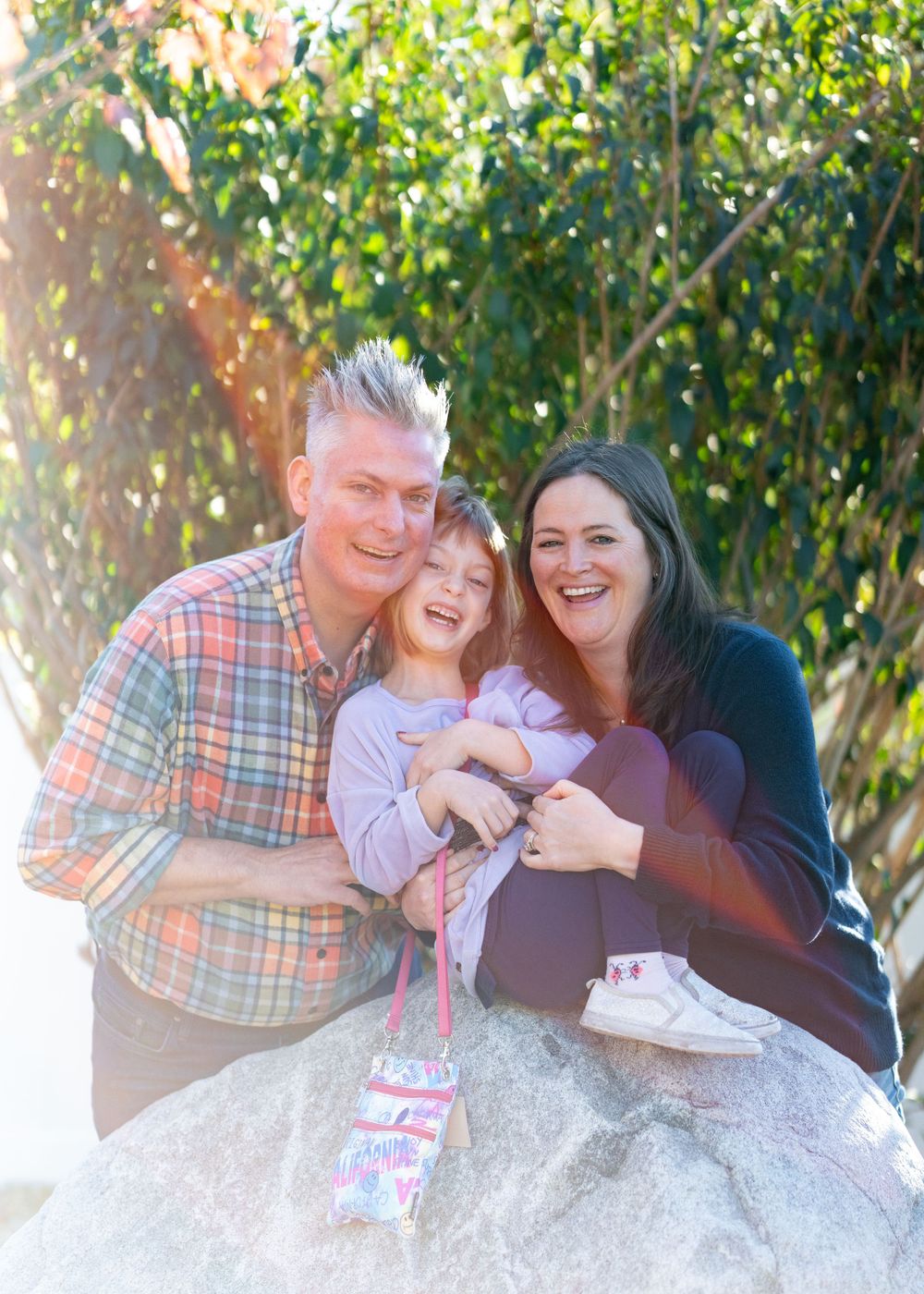 Family shares joyful moment together while sitting on large rock in sunlit backyard.