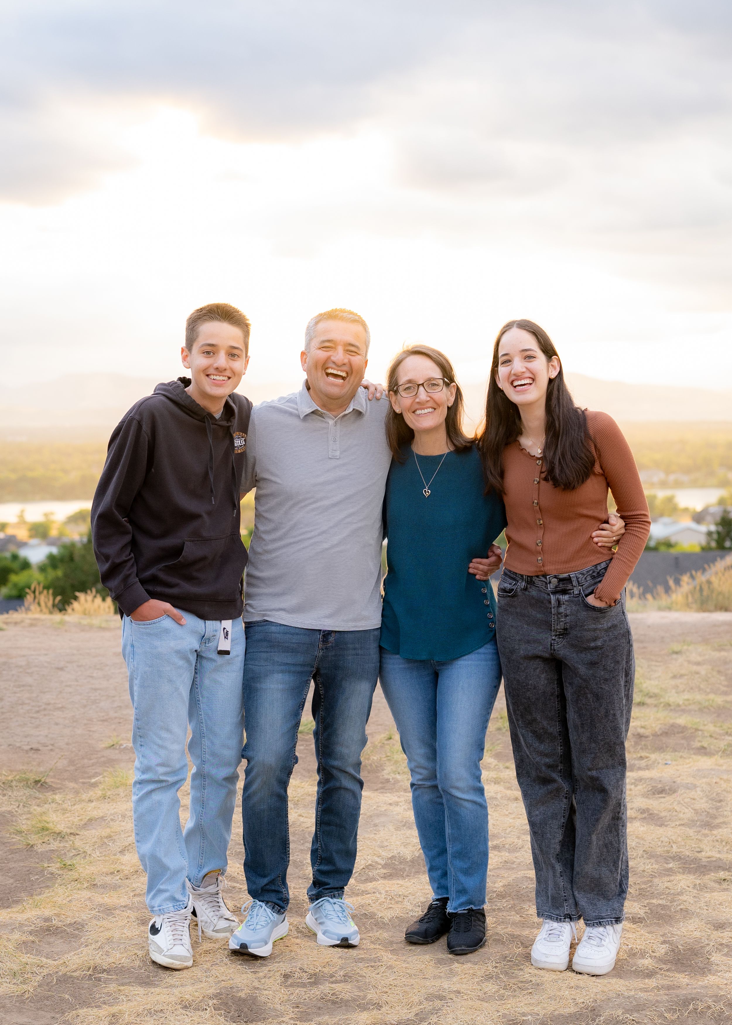 Family of four people stand close together smiling on a hilltop during sunset.