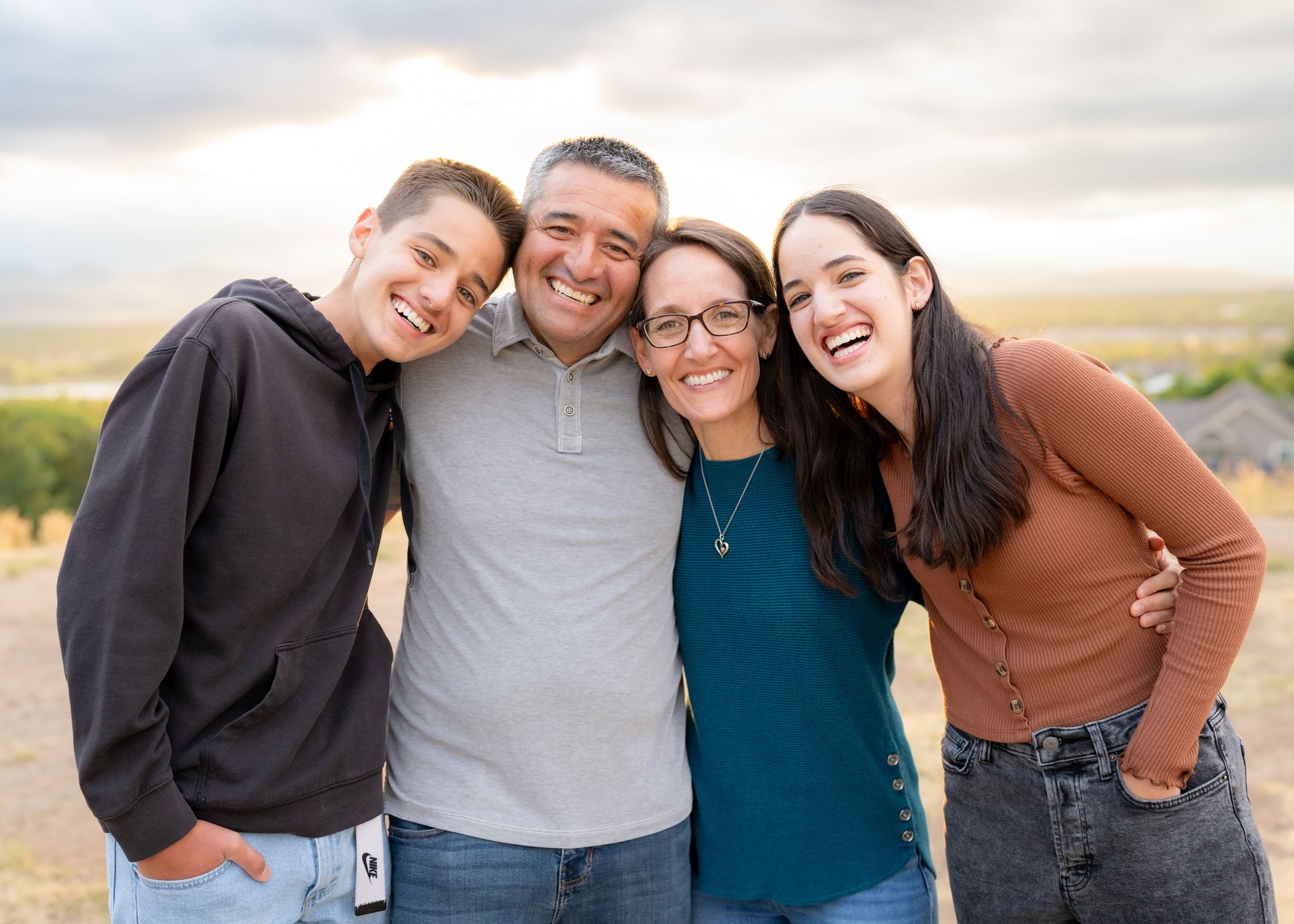 Family of four share laughter and embrace while posing for a casual outdoor photo at sunset.