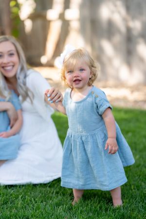 Toddler girl in blue dress laughing while playing on grass with her mother.