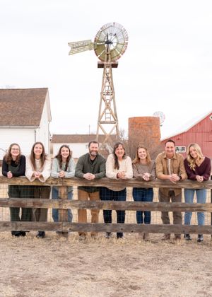 A group of people lean against a wooden fence at a rustic farm with a windmill and red barn in the background.
