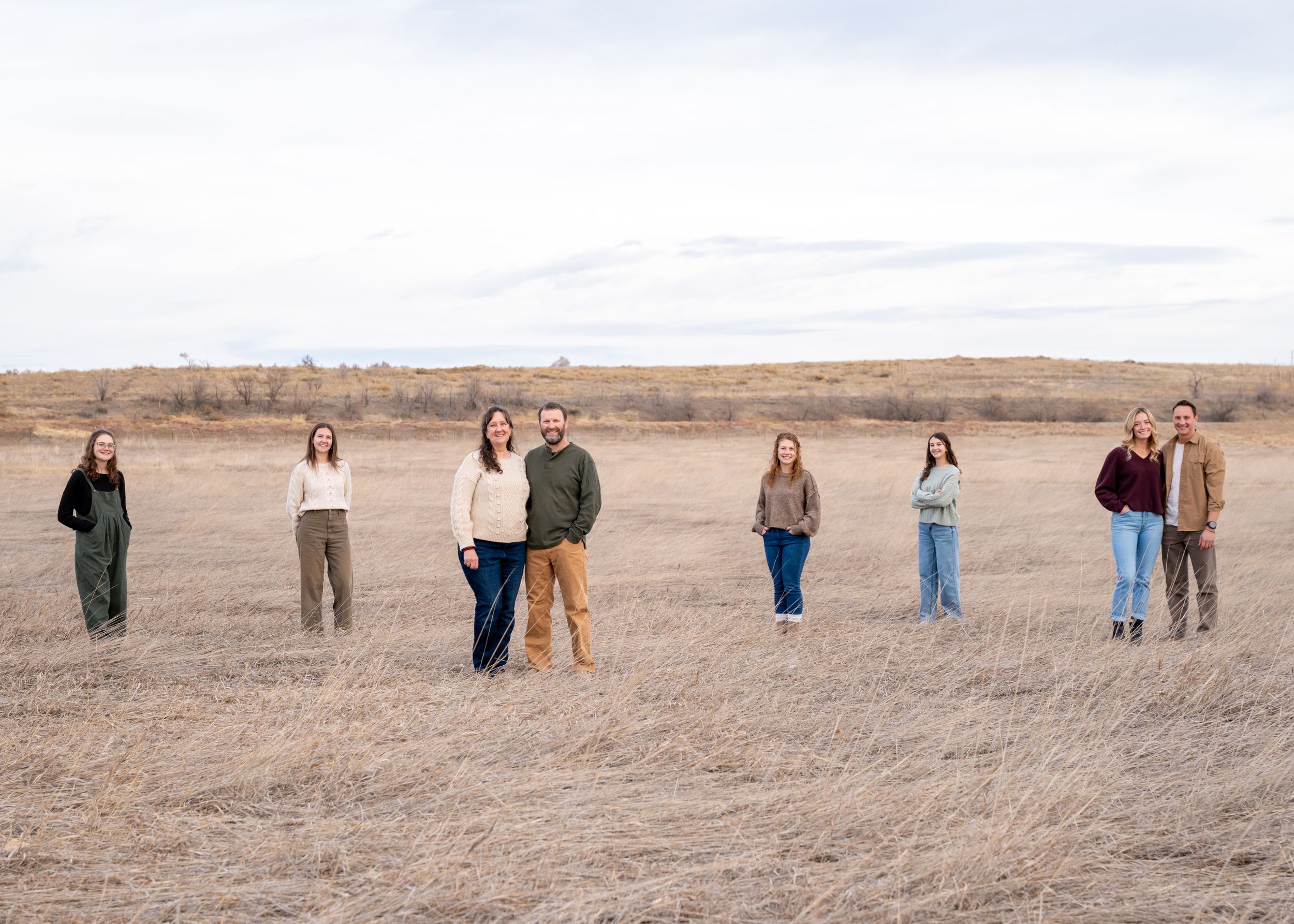 A group of friends stand spaced apart in a brown winter field under an overcast sky.