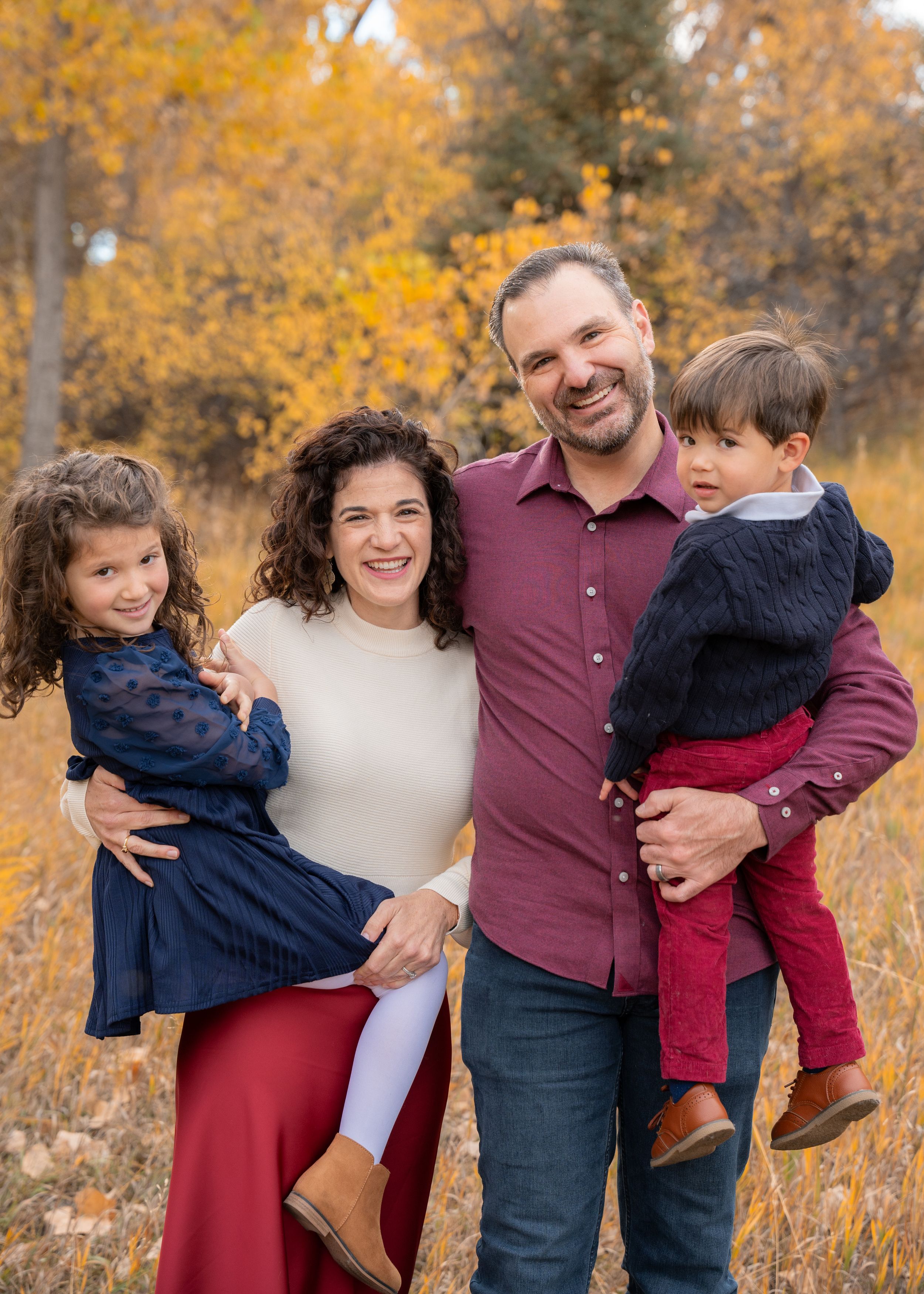 Family portrait outdoors with autumn colors and golden leaves in background.