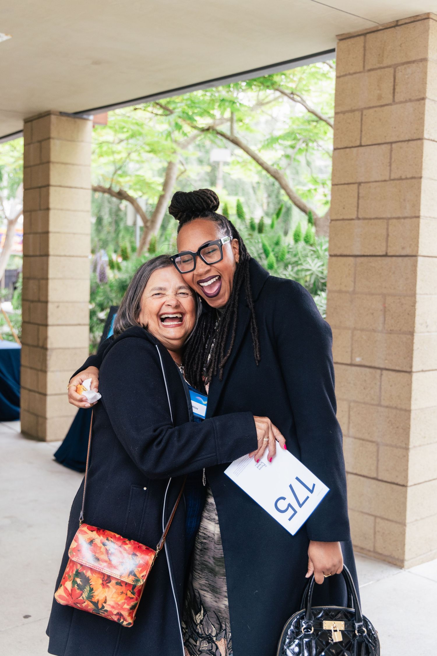 Two graduates in black caps and gowns share joyful embraces in front of pillars during a commencement ceremony.