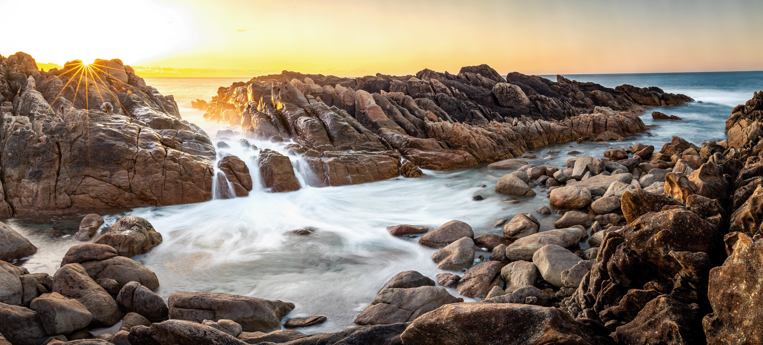 Long exposure seascape photograph of waves flowing through rocky coastal formations at sunset.