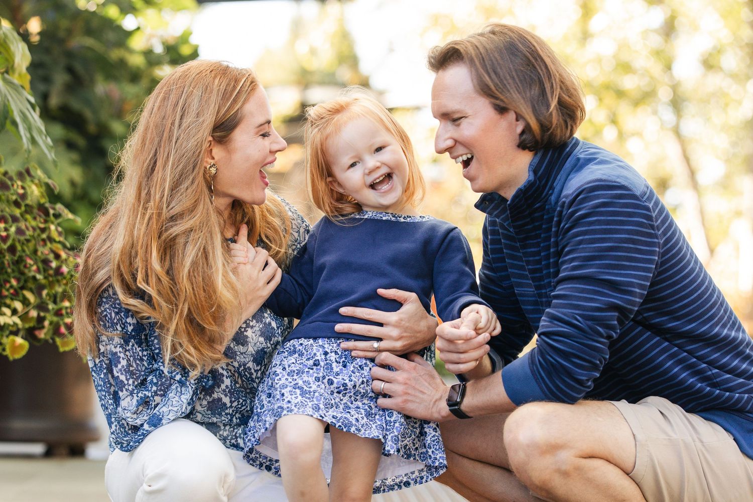 A joyful family moment outdoors with a laughing child in a blue dress being embraced by loved ones in casual wear.