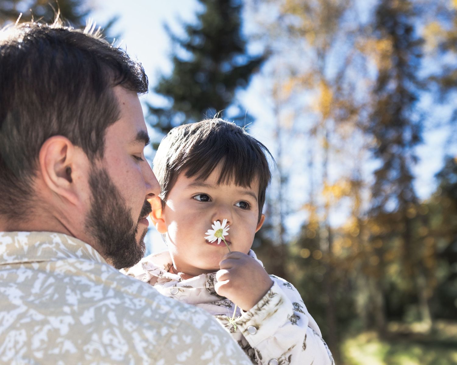Father with beard holds young toddler who holds a small white flower up to dad's nose outdoors.