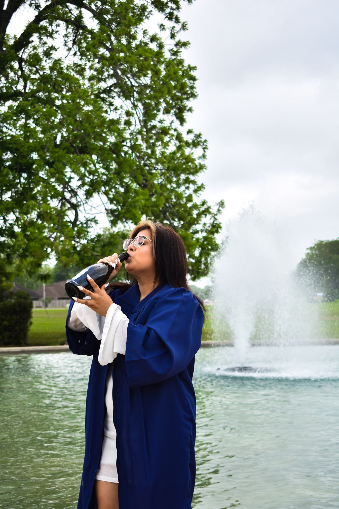 Graduate in blue commencement gown drinks from water bottle by fountain in park setting on cloudy day.