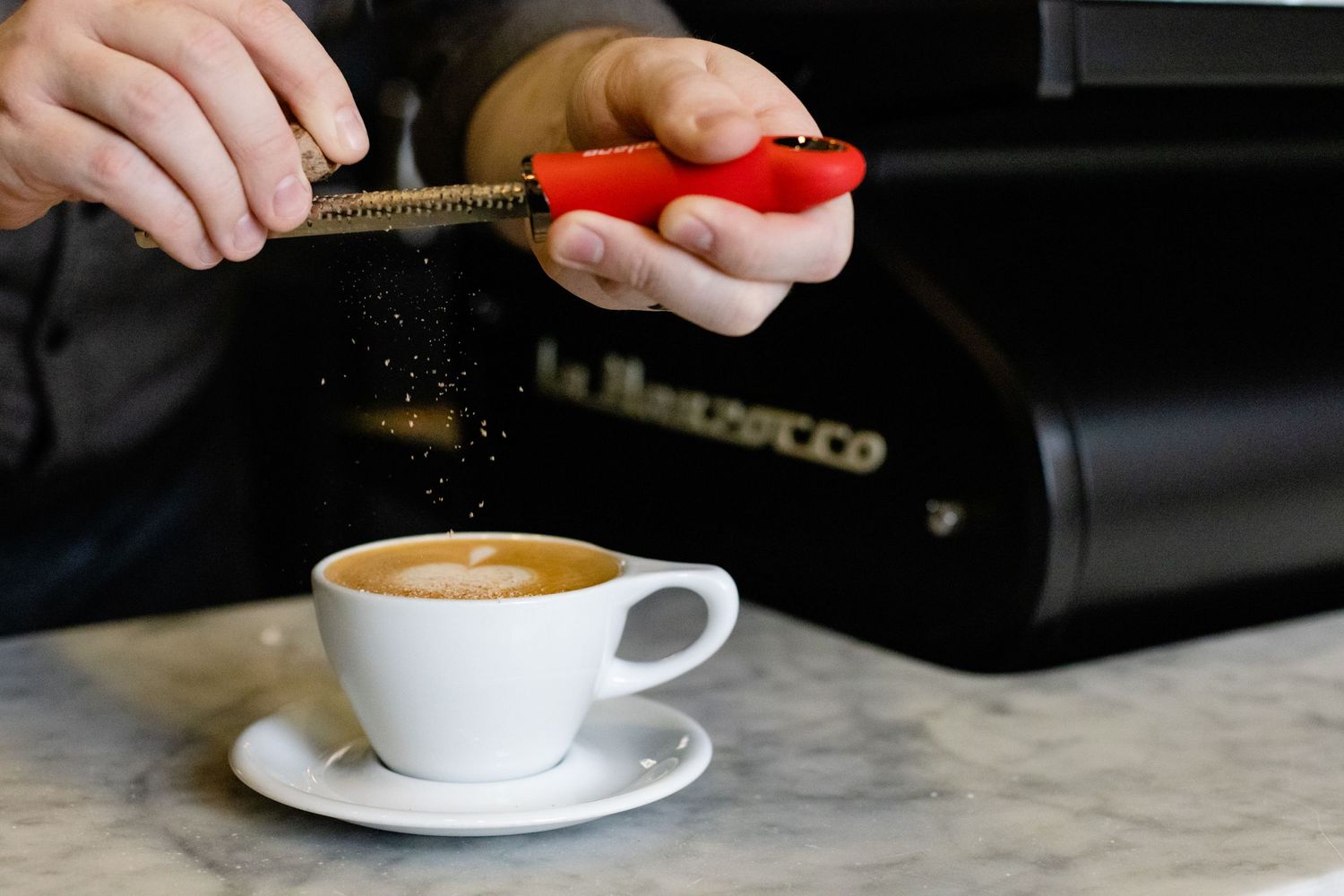 A coffee cup with latte art sits on a marble counter while someone uses a hand tamper above it.