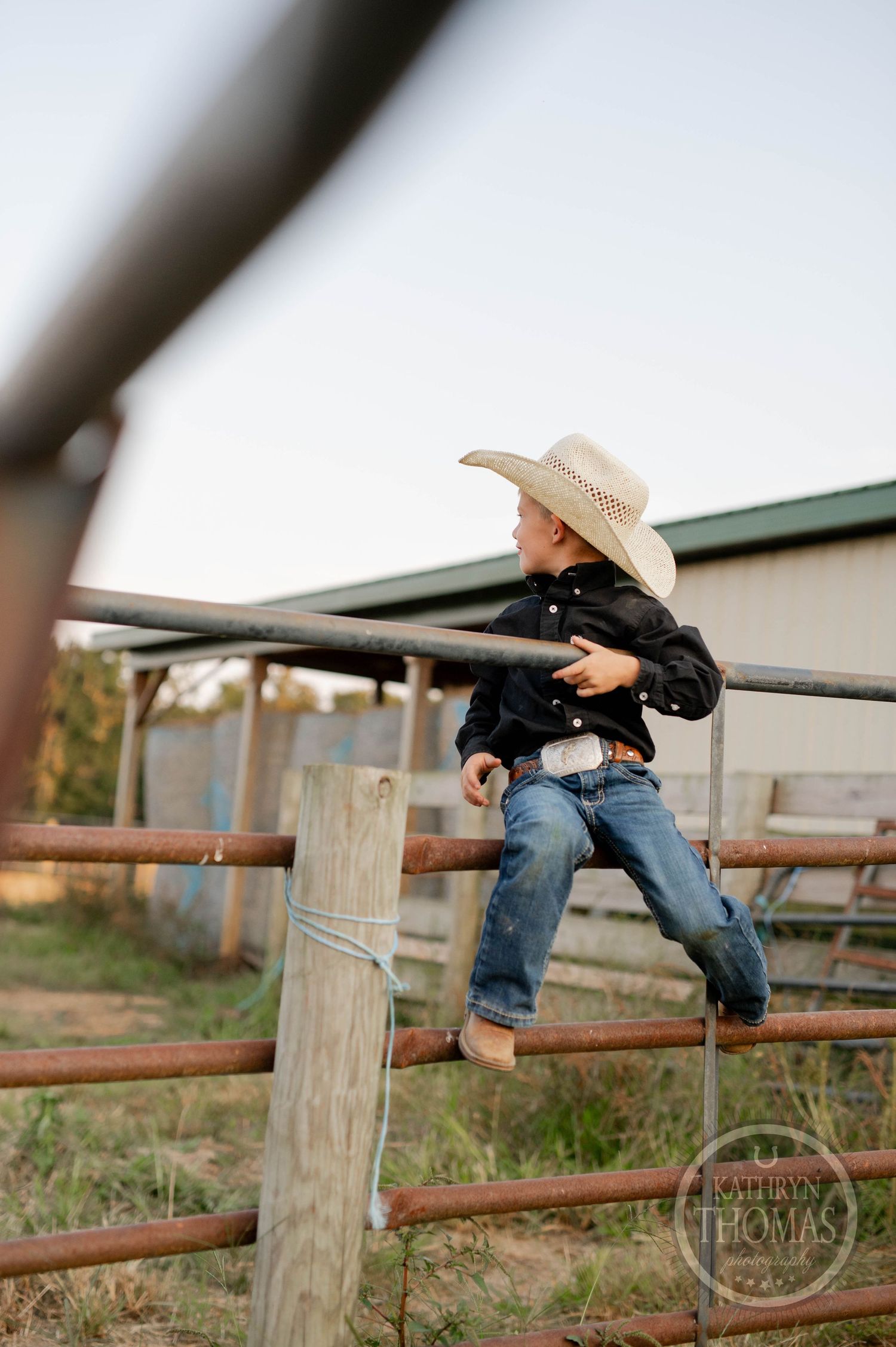 Child in western wear climbs metal fence rails at ranch during sunset.