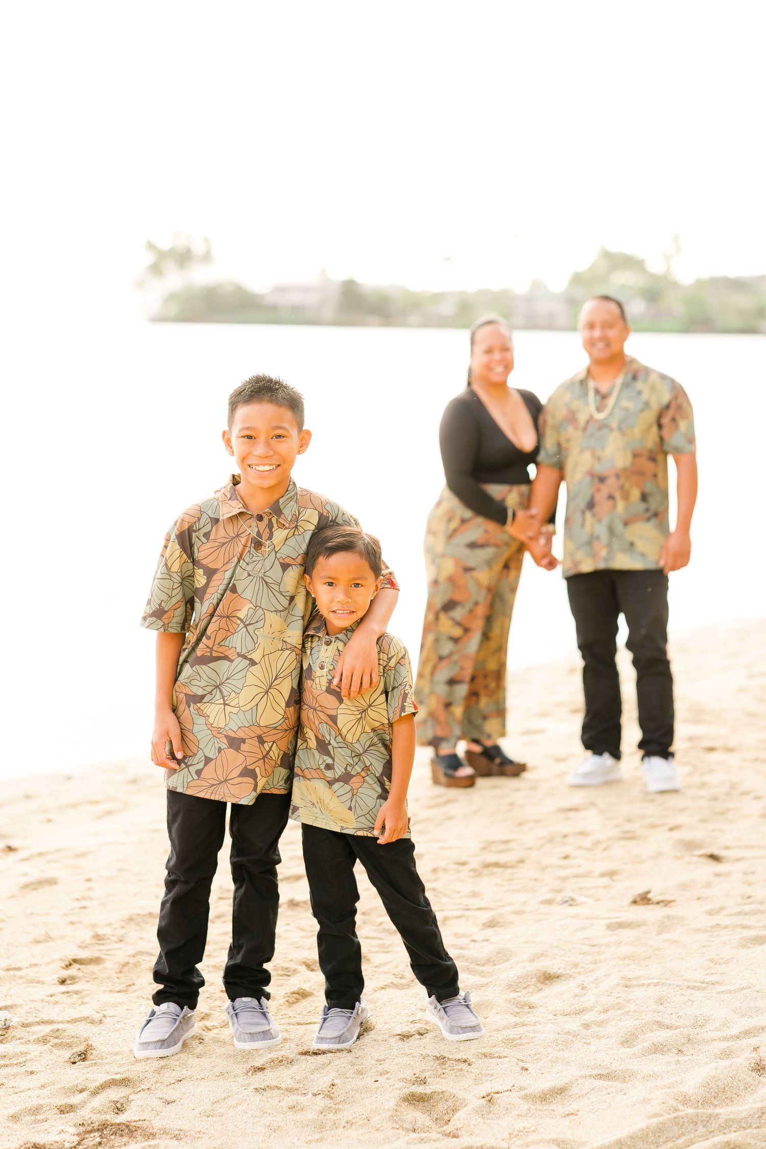 A family wearing coordinated Hawaiian print shirts poses together on a sandy beach at sunset.