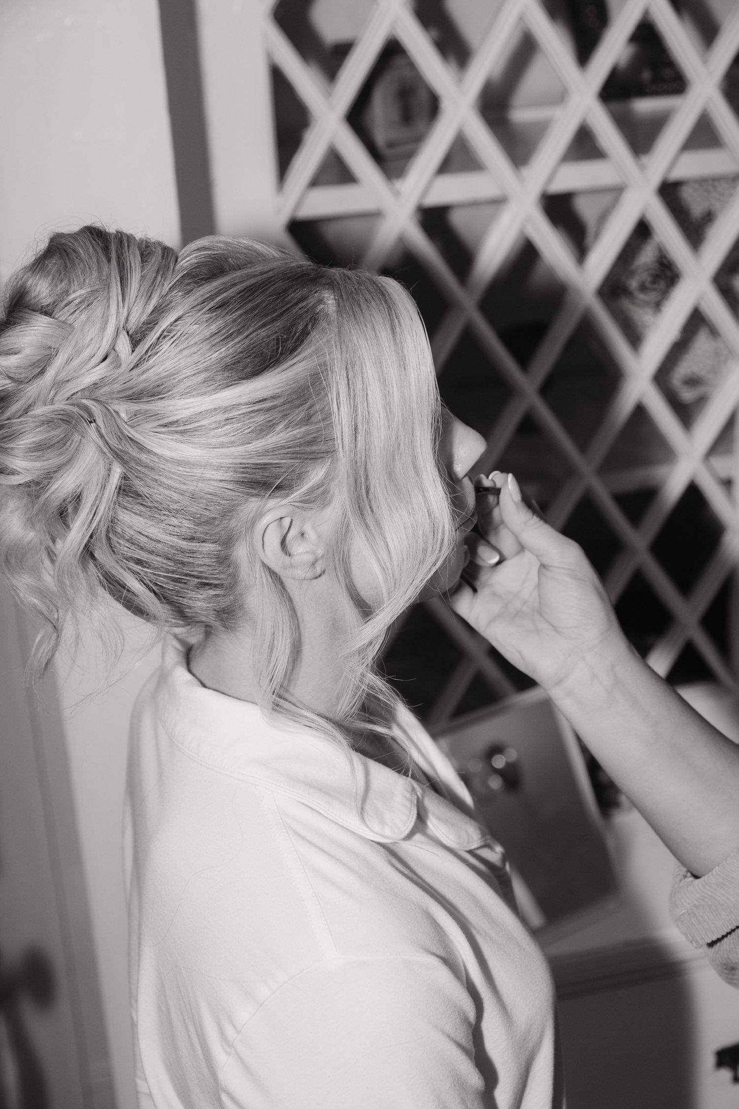 Blonde bride with elegant updo hairstyle getting makeup applied in black and white wedding preparation photo.