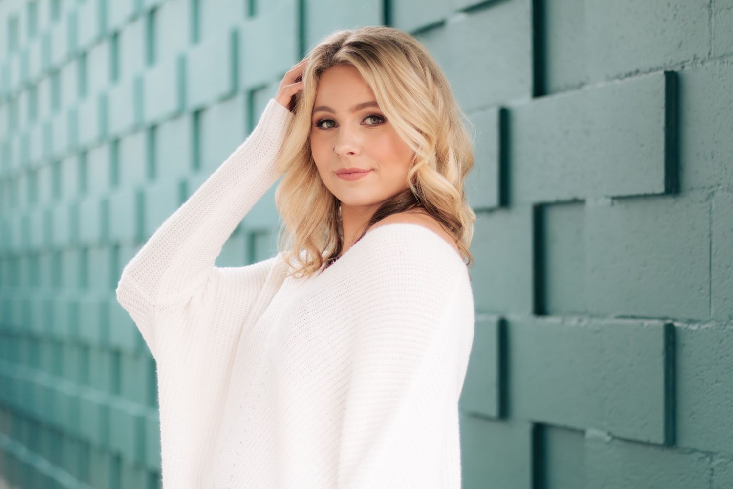 A person in a cozy white sweater poses against a teal brick wall with soft natural lighting and styled blonde hair.