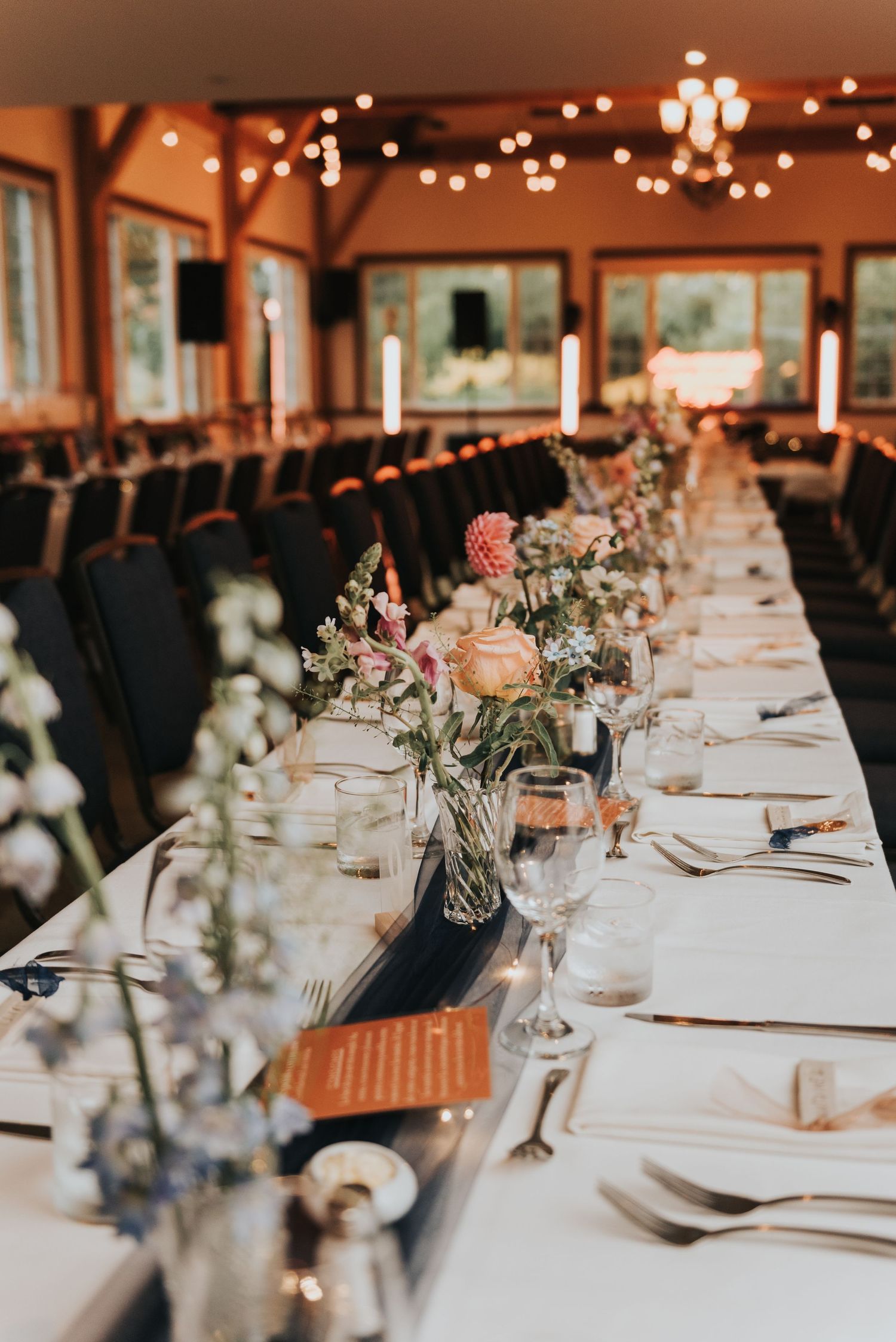 Long elegant wedding reception table set with white linens, black chairs and delicate floral centerpieces under warm lighting.
