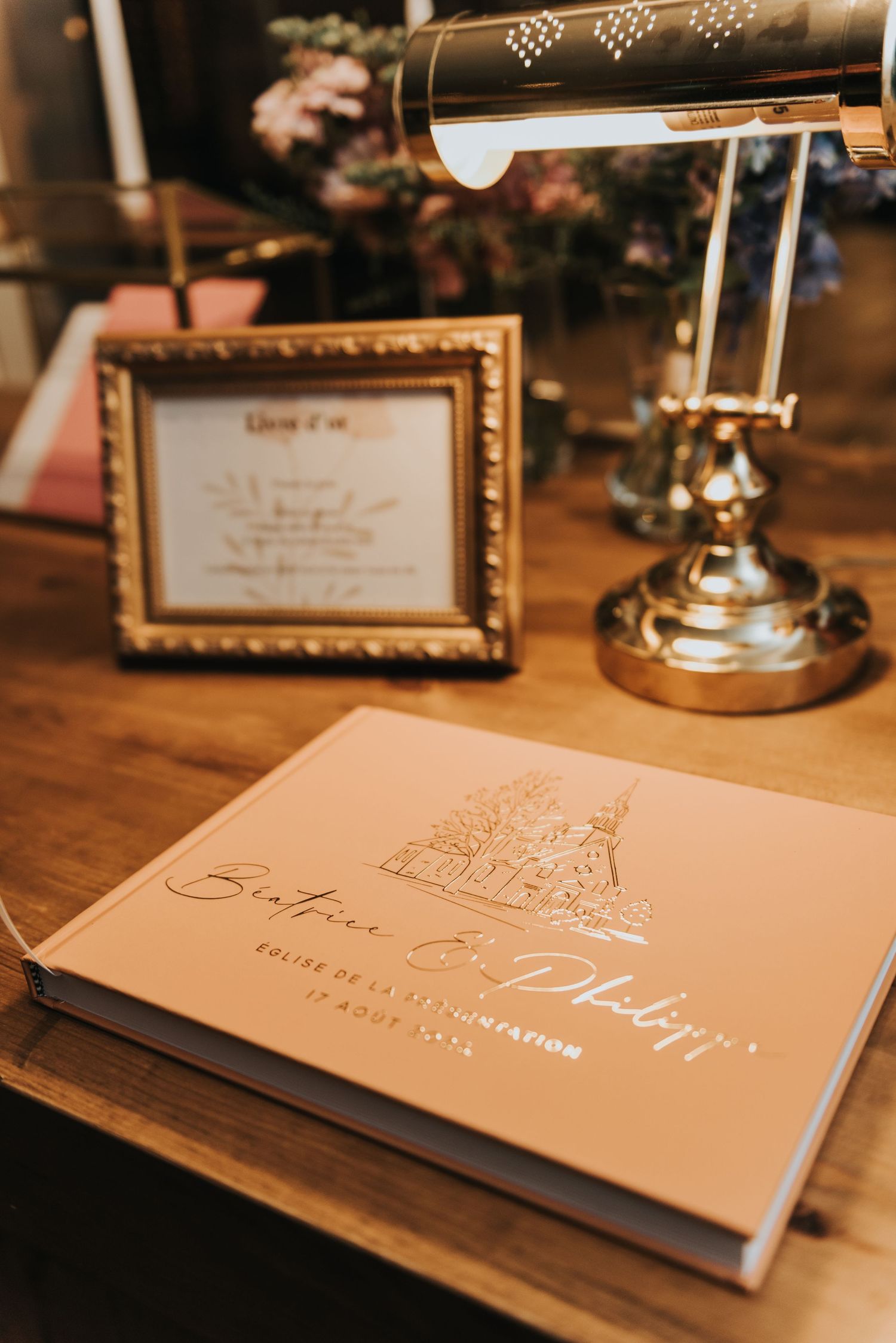 Gold-foiled wedding guest book with castle design sits on wooden desk next to framed sign and brass desk lamp.