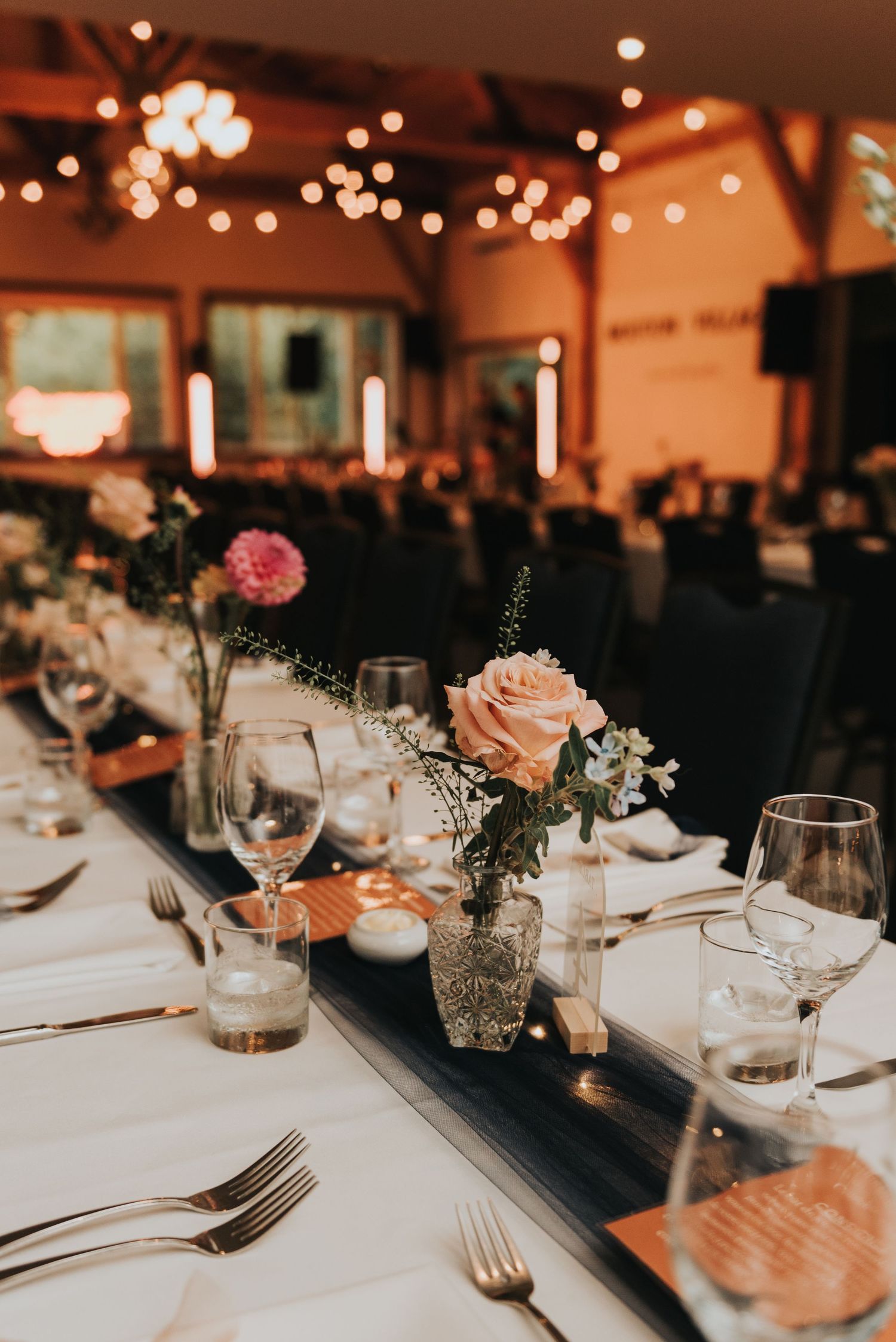Elegant wedding reception table setting with pink roses in vases, wine glasses and moody warm lighting.