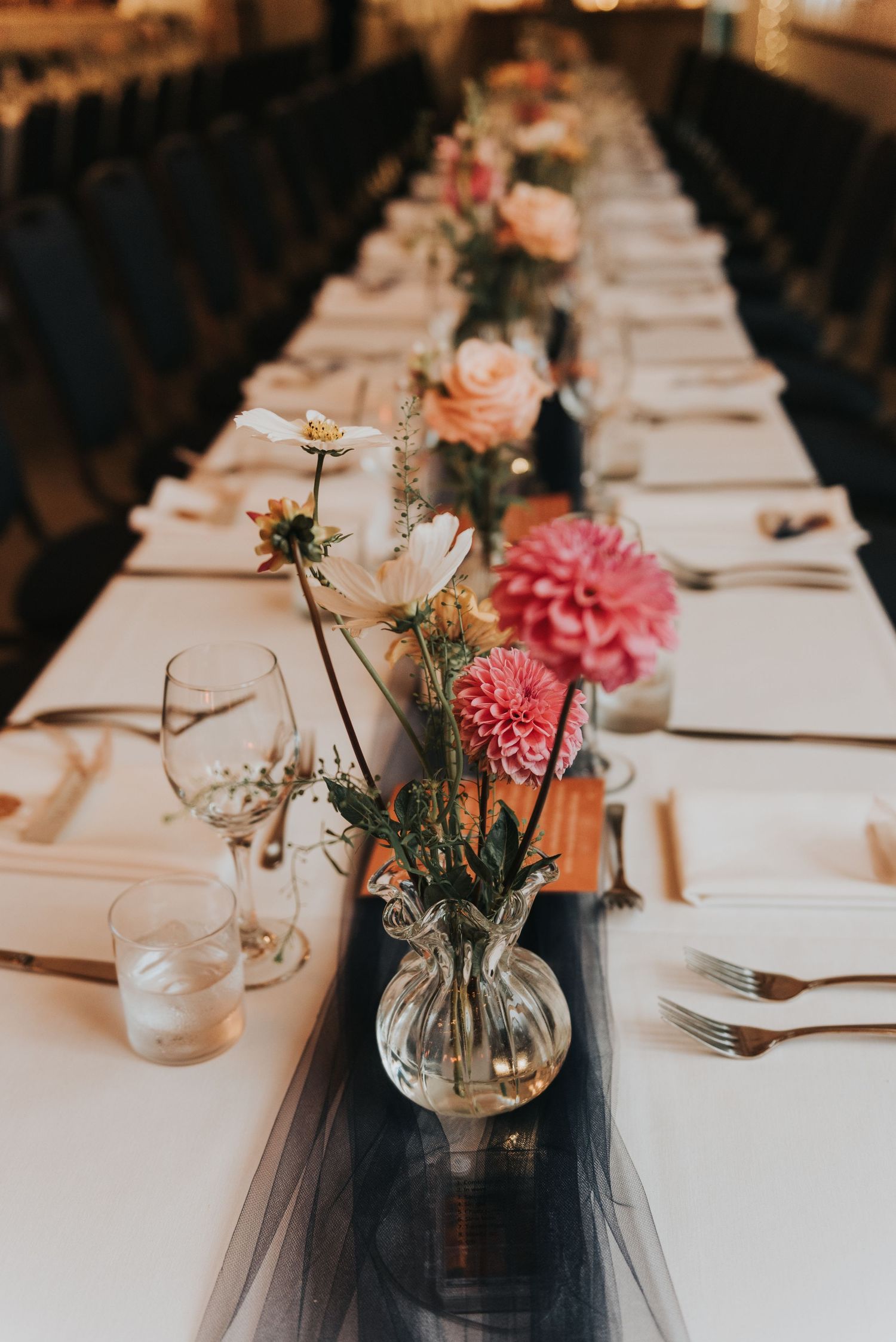 Close-up of wedding table decorations featuring pink dahlia flowers and roses in glass vases on black table runners.