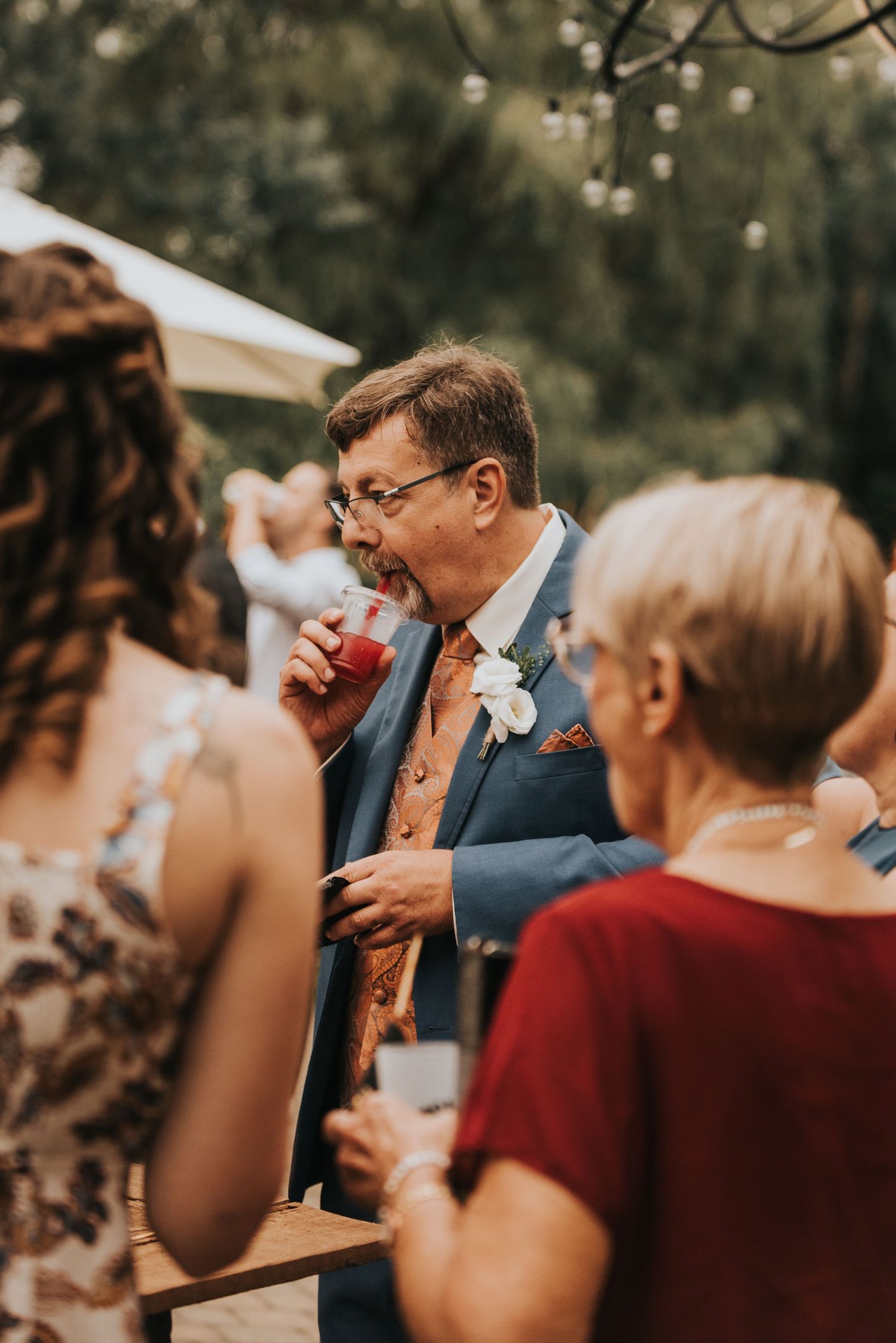 Wedding guests enjoy drinks and conversation at an outdoor reception under a white tent on a sunny day.