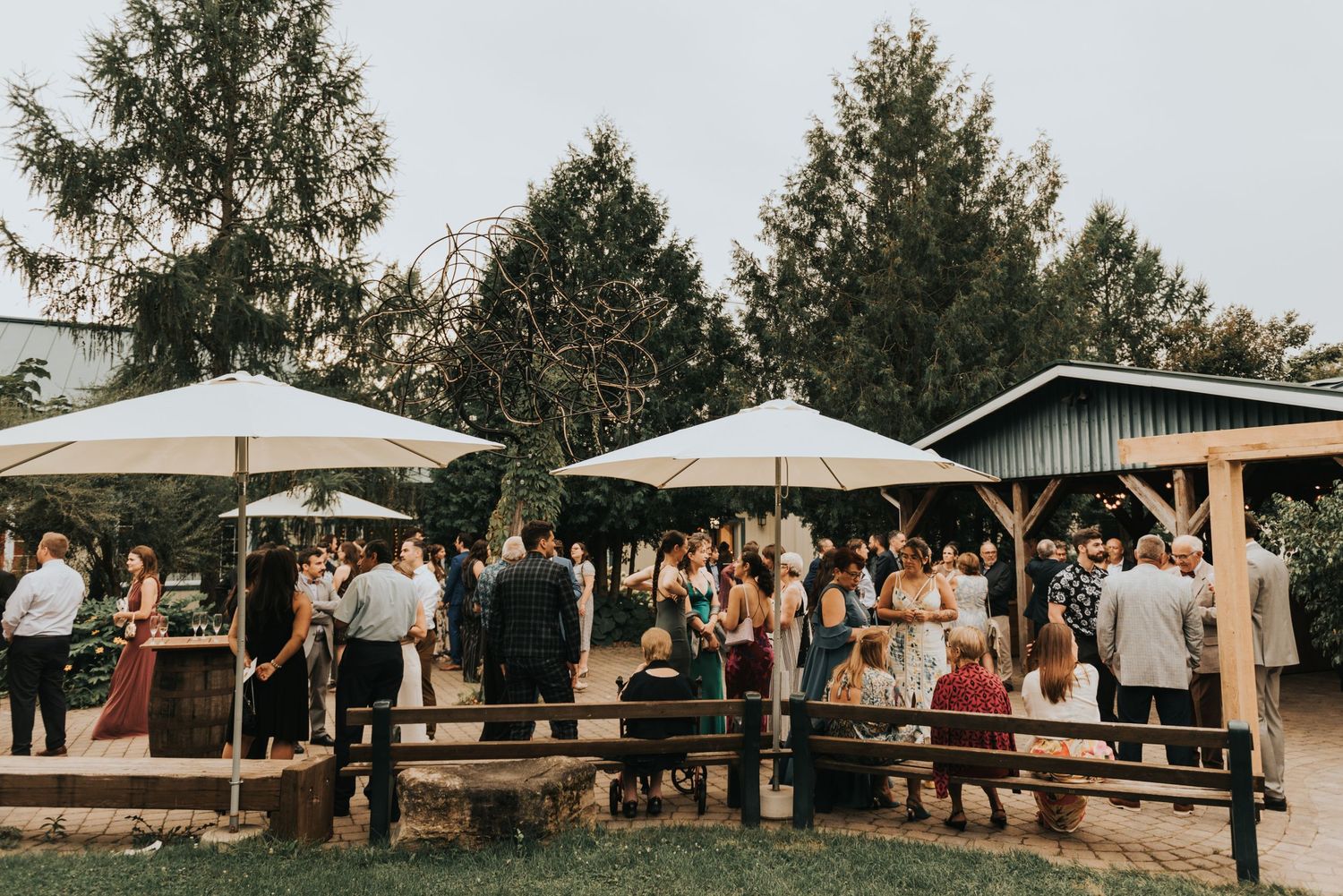 Wedding guests gather at an outdoor reception venue with white umbrellas and rustic wooden pavilions surrounded by evergreen trees.