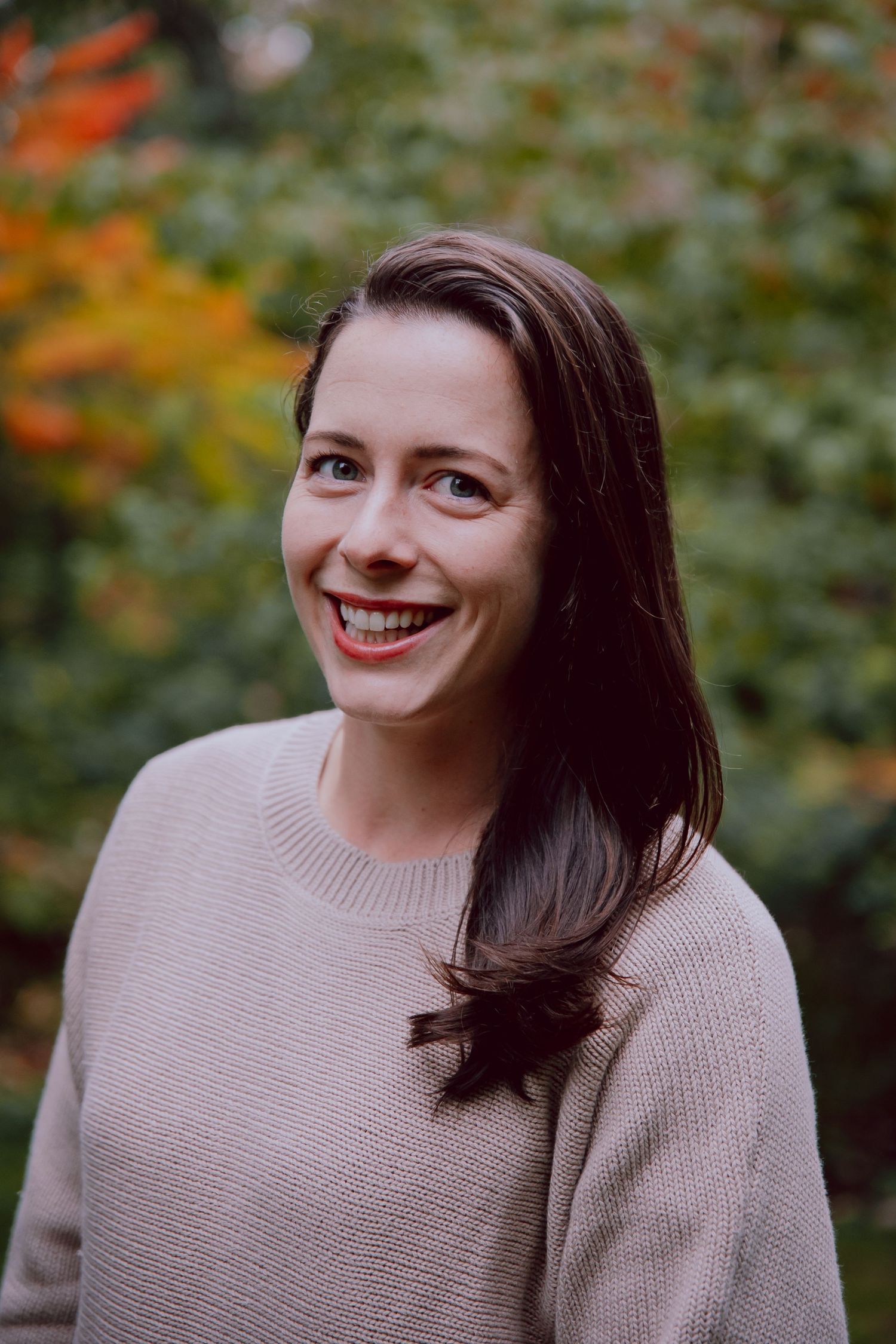 A woman in a beige sweater smiles warmly at the camera in an autumn outdoor setting.